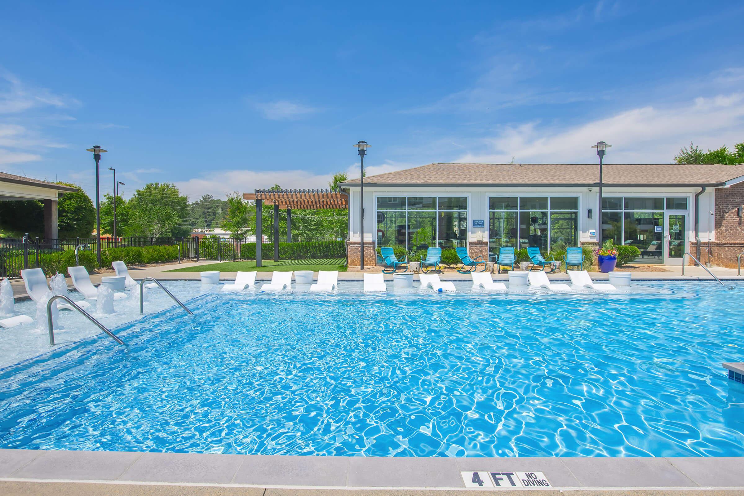 A bright outdoor swimming pool with clear blue water, surrounded by lounge chairs. In the background, there is a building with large windows and a pergola. The scene is set in a sunny environment with green trees and blue skies, creating a relaxing atmosphere. A "4 FT NO DIVING" sign is visible near the pool edge.