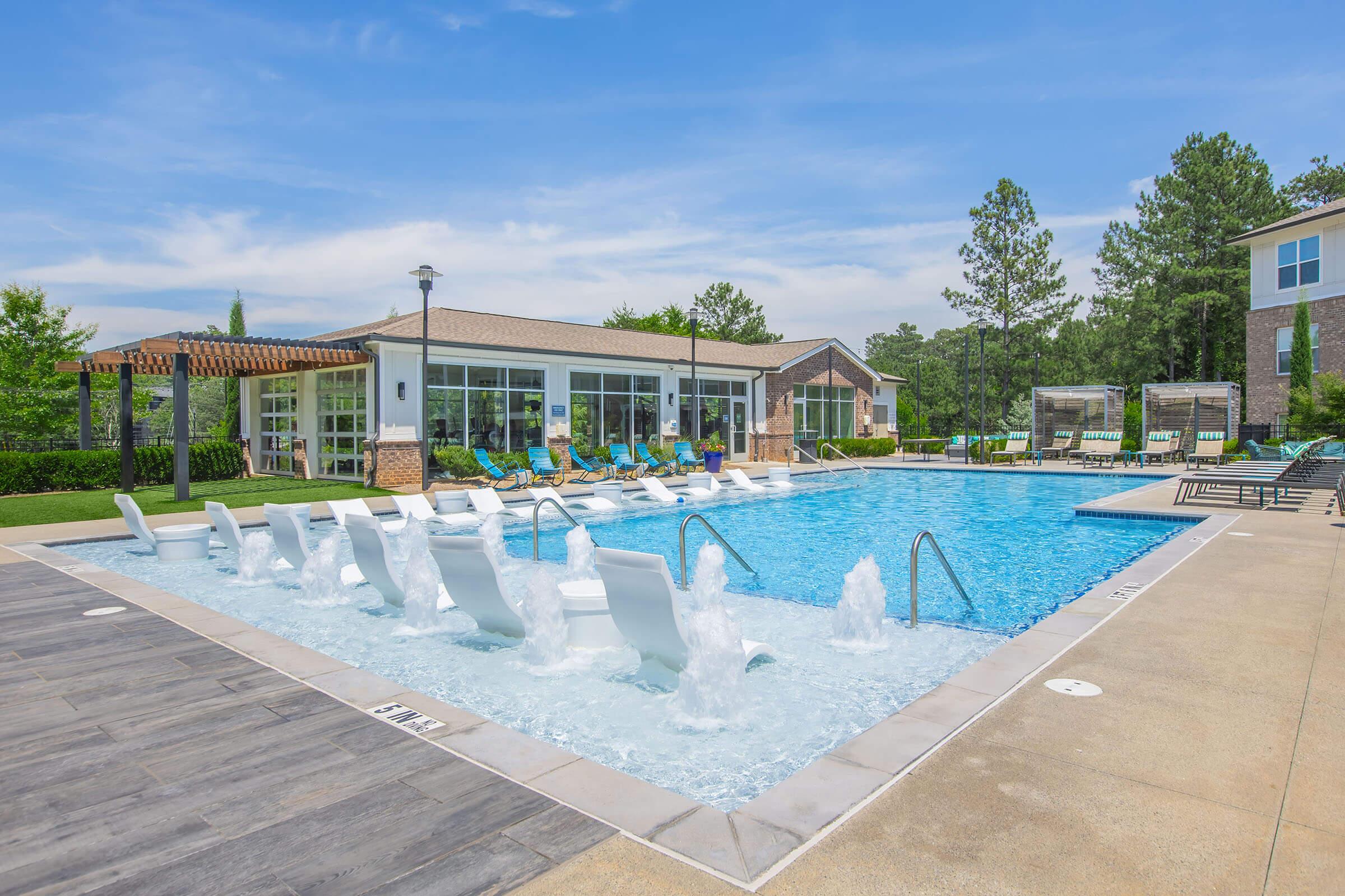 A sparkling outdoor swimming pool with lounge chairs partially submerged in the water, surrounded by greenery and modern seating areas. Sunlight shines on the pool deck, creating a relaxing atmosphere, with a clubhouse visible in the background.