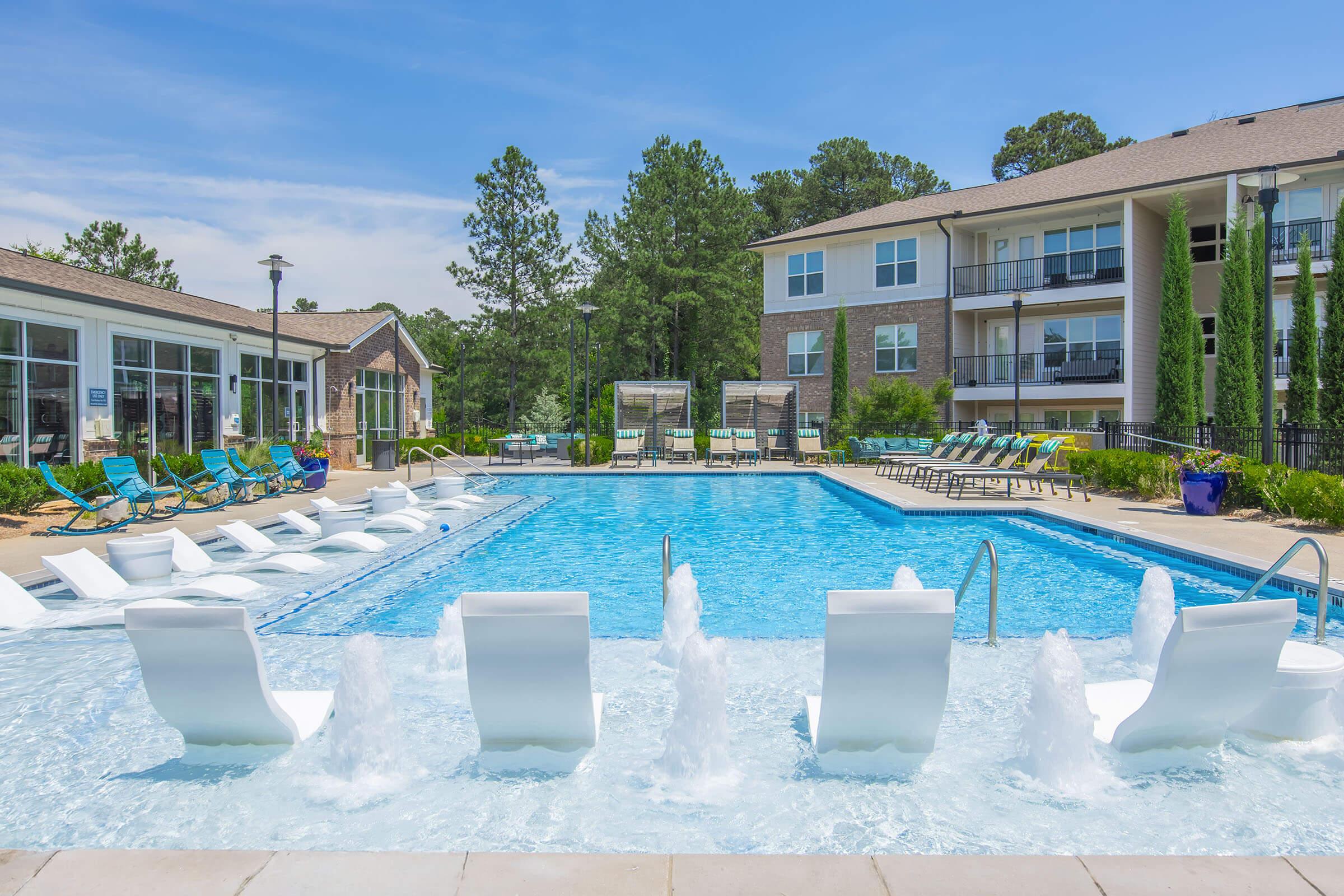 A spacious outdoor swimming pool surrounded by lounge chairs, with water features creating a relaxing atmosphere. The pool area is bordered by well-maintained greenery and modern apartment buildings in the background. Bright blue sky adds to the inviting ambiance.