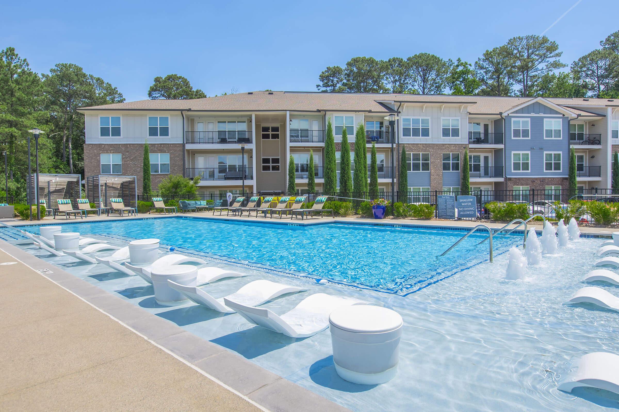 A vibrant outdoor pool area surrounded by modern apartment buildings. The pool features lounge chairs, a decorative fountain, and lush greenery. Clear blue skies overhead create a sunny, inviting atmosphere perfect for relaxation and leisure.