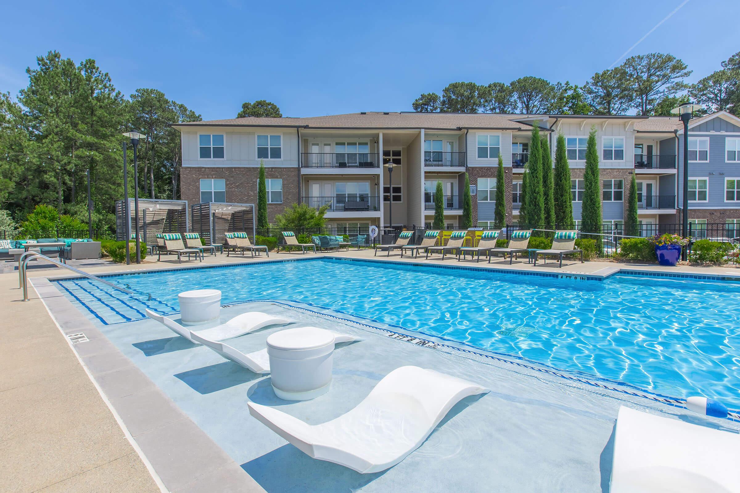 A bright outdoor swimming pool area featuring lounge chairs along the water, surrounded by landscaped greenery. In the background, there are modern apartment buildings with balconies. The sky is clear and blue, creating a welcoming atmosphere for relaxation and leisure.