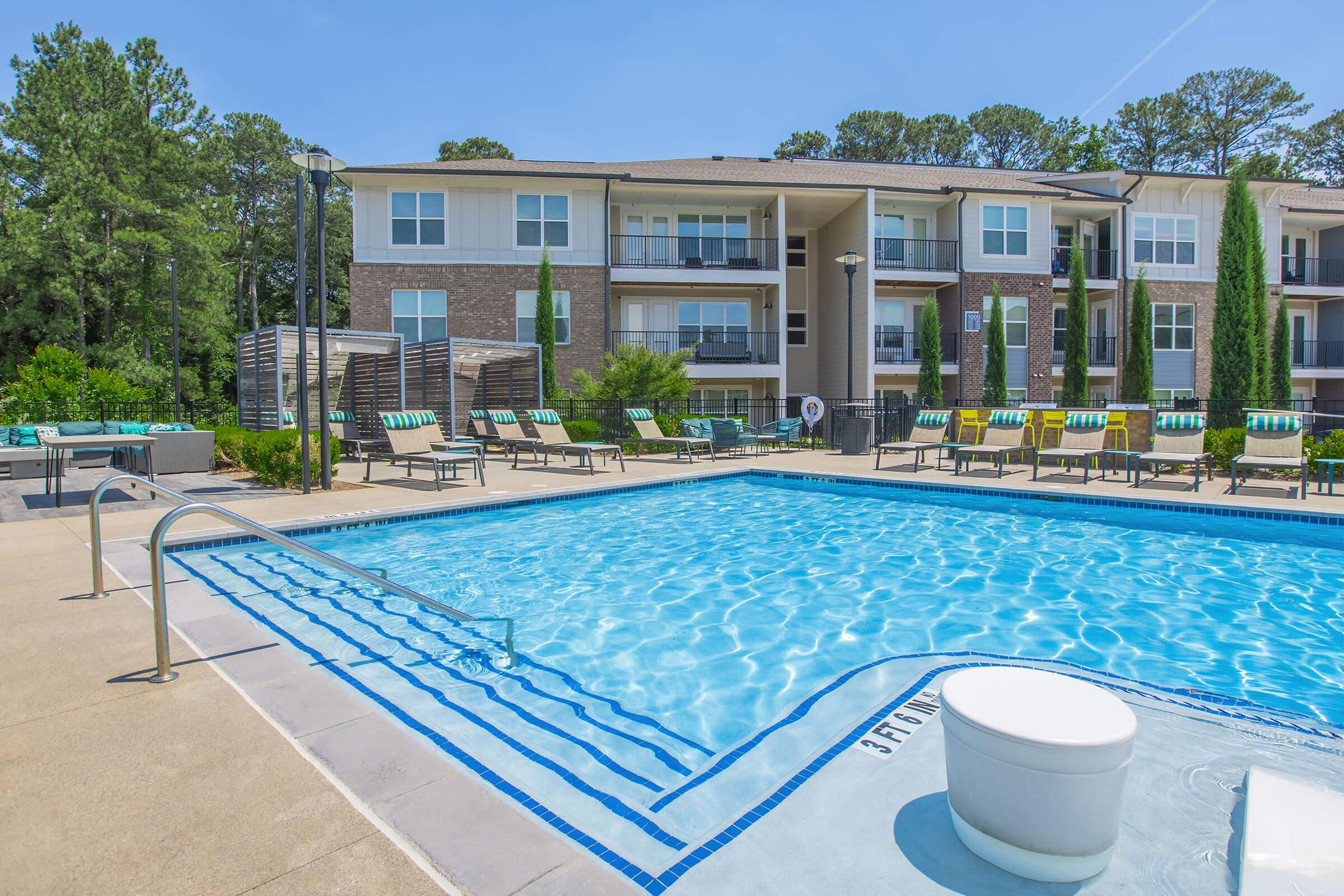 A clear swimming pool with blue water, surrounded by lounge chairs and shaded areas. In the background, a modern apartment building with balconies overlooks the pool area. Lush green trees line the property, creating a serene outdoor space. Bright sunny day with clear skies.