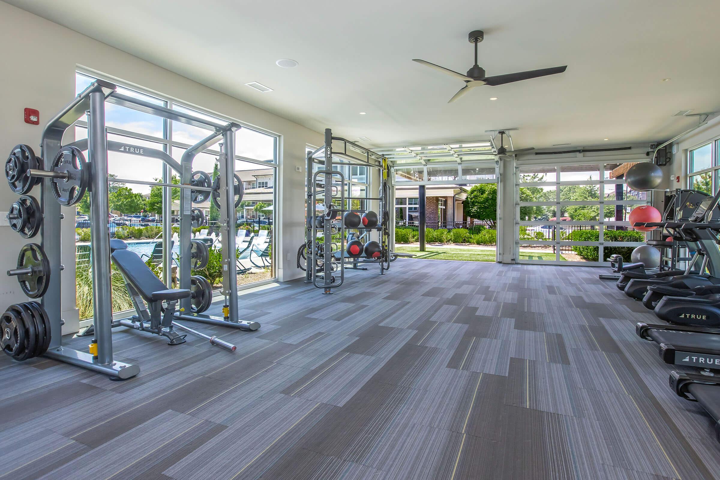 A modern gym interior featuring weightlifting equipment, exercise mats, and large windows overlooking a pool area. The space is bright and airy, with a fan on the ceiling, showcasing a clean and organized layout ideal for workouts.