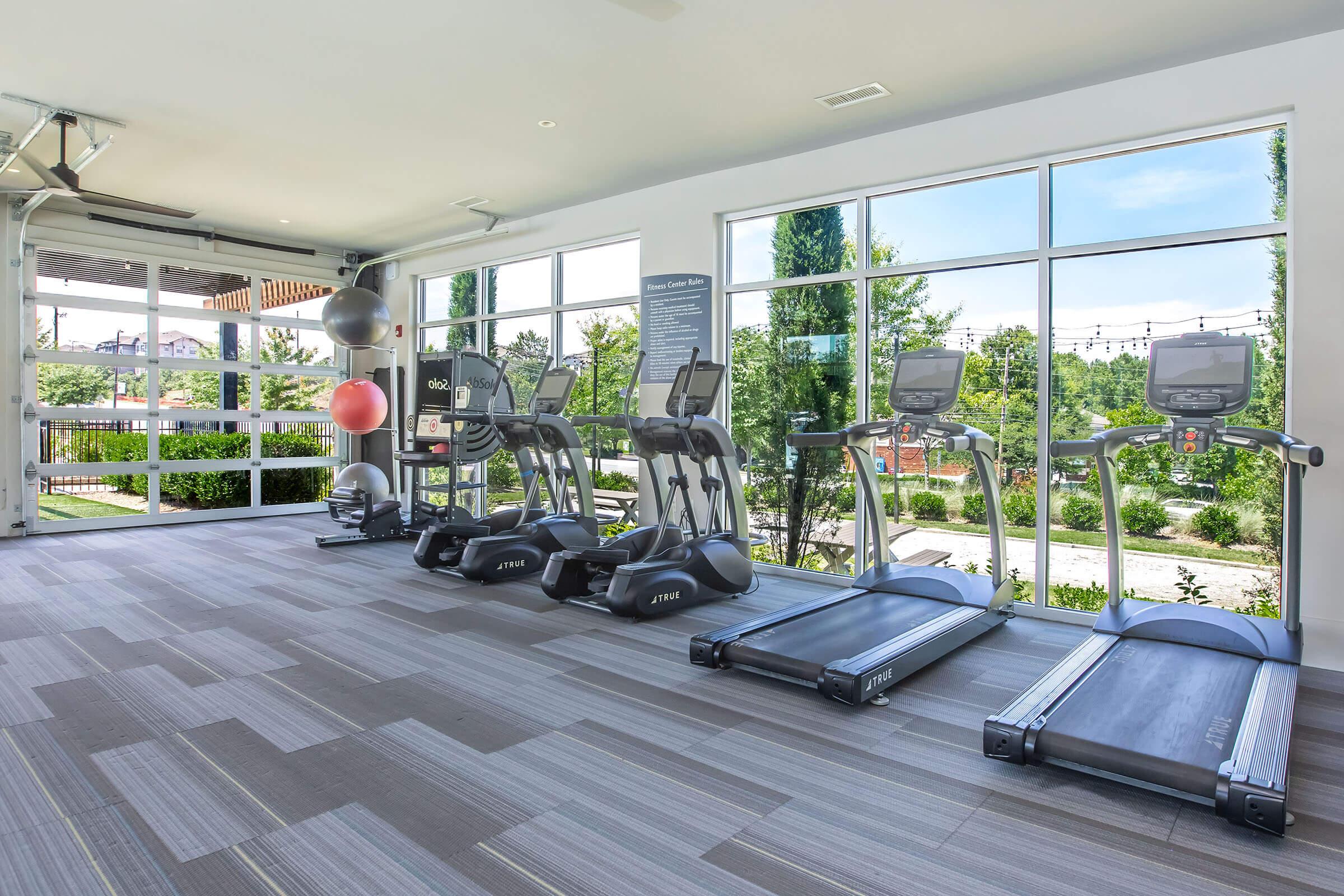 A modern gym interior featuring several treadmill machines and elliptical trainers lined up against large windows. The space is bright and open, with green trees and outdoor seating visible outside. The flooring is gray with a contemporary design, and there are fitness accessories like stability balls in the area.