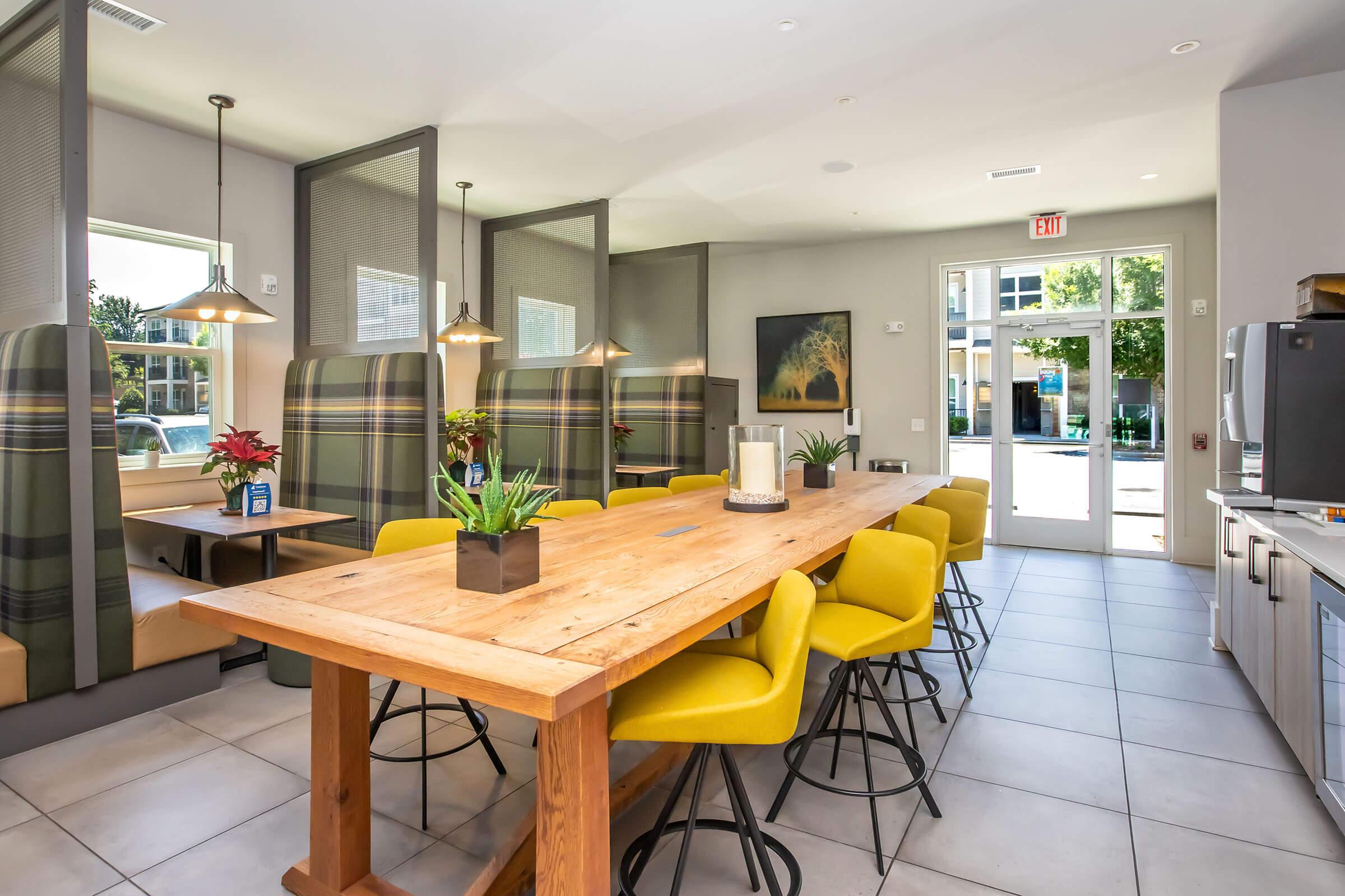 Modern dining area featuring a large wooden table surrounded by bright yellow chairs. Booth seating with plaid upholstery lines the sides, and large windows allow natural light to fill the space. Decorative plants and artwork add a touch of warmth to the contemporary design.