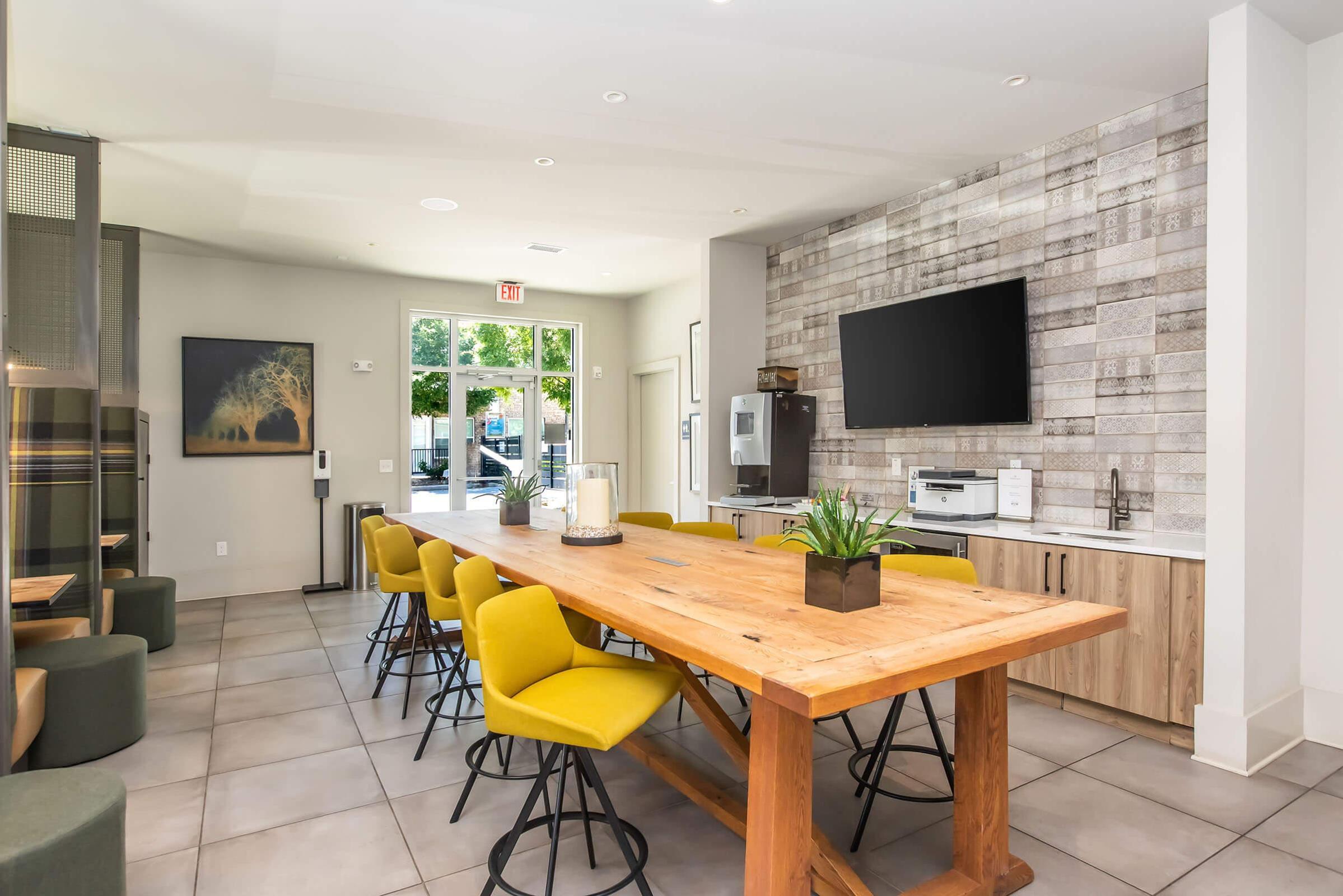 A modern kitchen or dining area featuring a large wooden table surrounded by yellow chairs. The space includes a flat-screen TV mounted on the wall and a small kitchenette with a microwave and other appliances. Large windows provide natural light, and there are green accents in the decor.