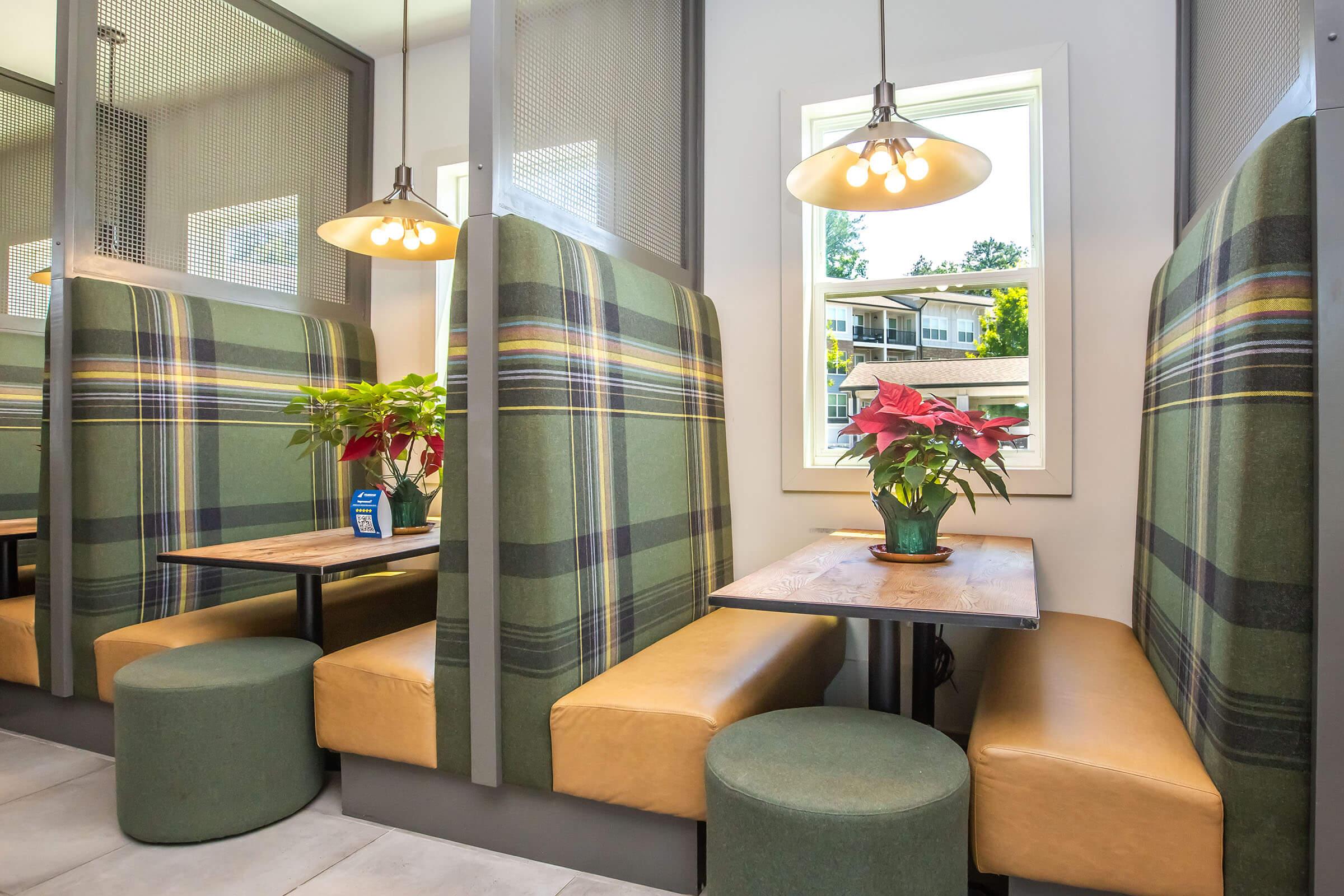 Cozy restaurant seating area with green plaid booths and round green stools. Each booth has a small wooden table and a decorative potted plant in the center. Large windows allow natural light to fill the space, creating a warm and inviting atmosphere.