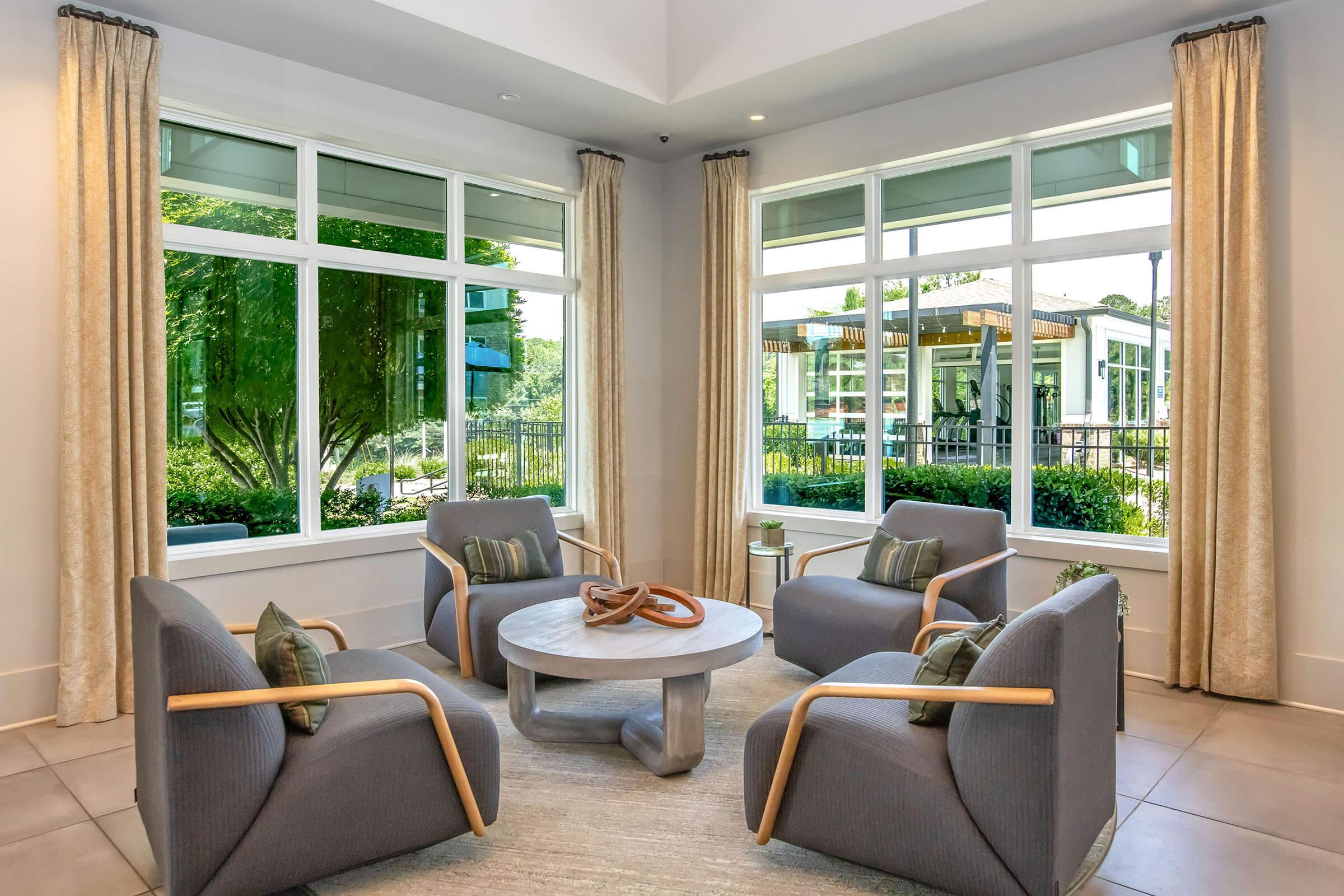 A cozy lounge area with four modern gray chairs and a round table in the center. Large windows with light curtains let in natural light, revealing outdoor greenery and poolside views. The space is designed for relaxation and socializing.