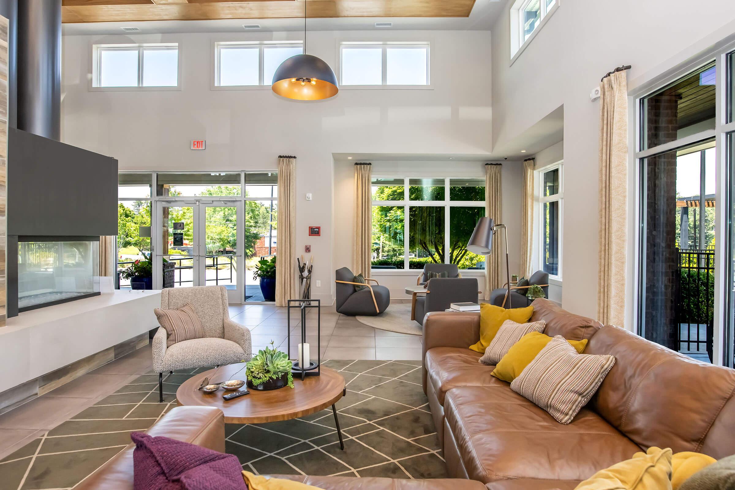 Cozy living room area featuring a large brown leather sofa with yellow accent pillows, a round coffee table with plants, and modern decor. Bright natural light streams in through large windows, showcasing a view of greenery outside. A sleek fireplace and contemporary lighting add to the inviting atmosphere.