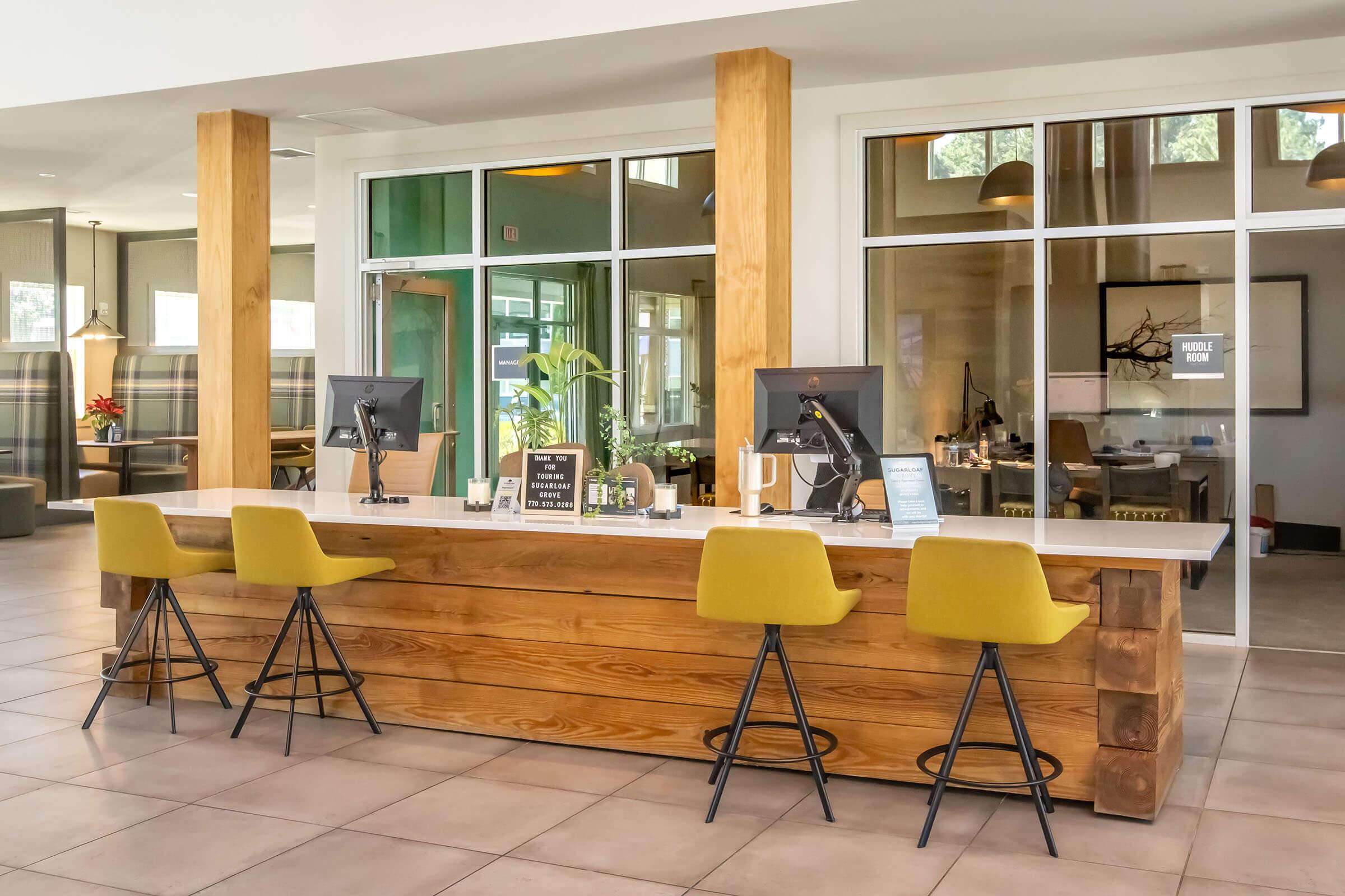 A modern reception area featuring a wooden front desk with two computer stations. Four yellow bar stools are positioned in front of the desk. The space has large glass windows allowing natural light in, and decorative plants add a fresh touch. Soft lighting creates a welcoming atmosphere.