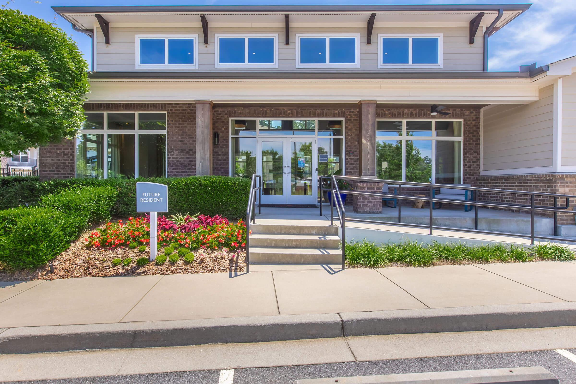 Exterior view of a modern building with large windows and a welcoming entrance. The path features a ramp for accessibility leading to the main door. Colorful landscaping with flowers is present in front, and a sign labeled "FUTURE RESIDENT" is positioned near the walkway. Clear blue sky forms the backdrop.