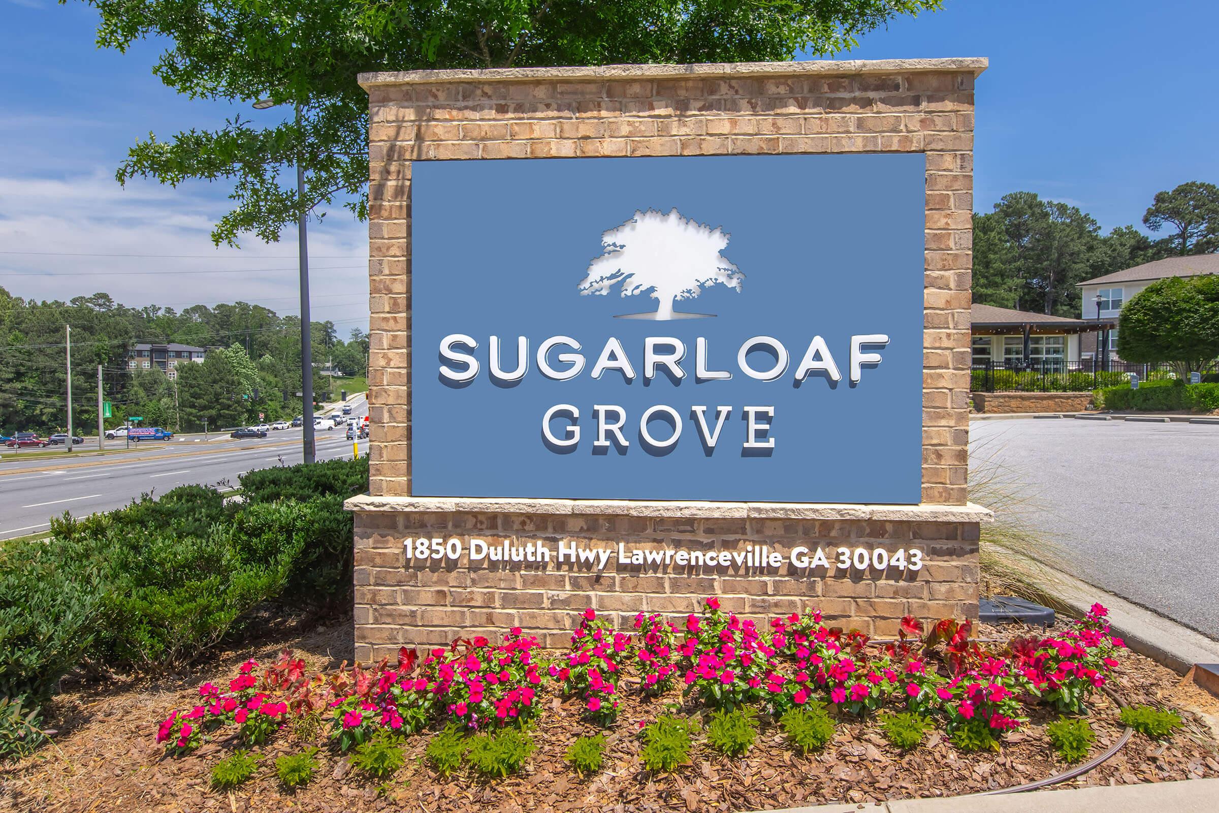 Sign for Sugarloaf Grove located at 1850 Duluth Hwy, Lawrenceville, GA 30043. The sign features a large tree logo and is surrounded by colorful flower beds and greenery, set against a blue sky and a clear view of the road.