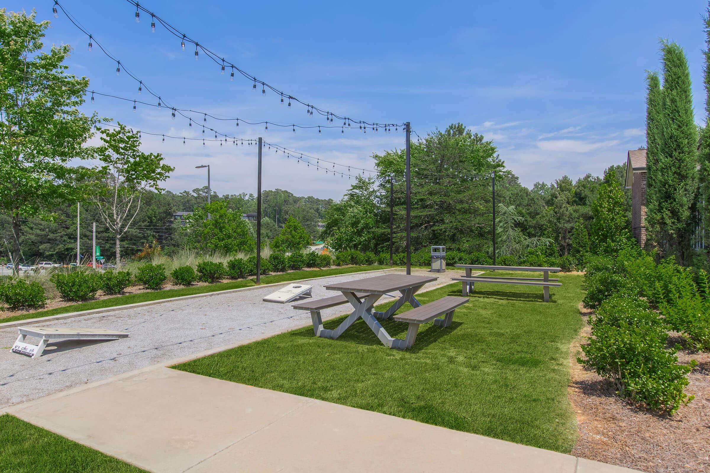 A well-kept outdoor area featuring picnic tables on a grassy patch, string lights overhead, and a gravel surface for games. Lush greenery and trees surround the space, providing a pleasant environment for relaxation and social gatherings.