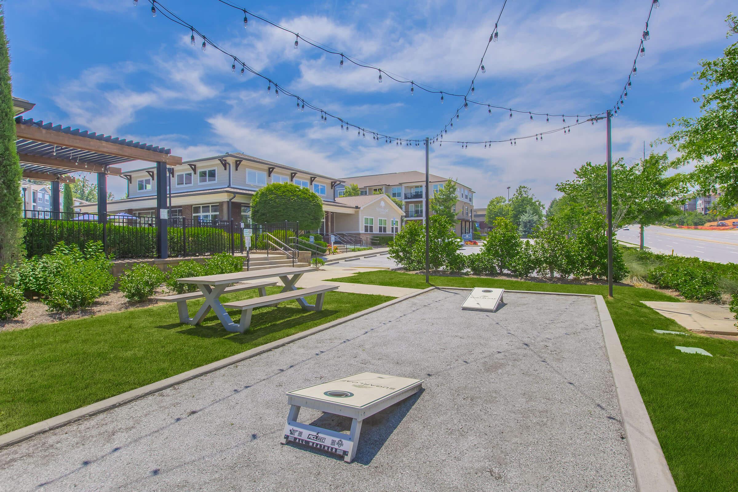 A sunny outdoor area featuring a cornhole game setup on a gravel surface, surrounded by green grass and landscaping. There are picnic tables nearby, string lights overhead, and residential buildings in the background. The scene conveys a relaxing and social environment.