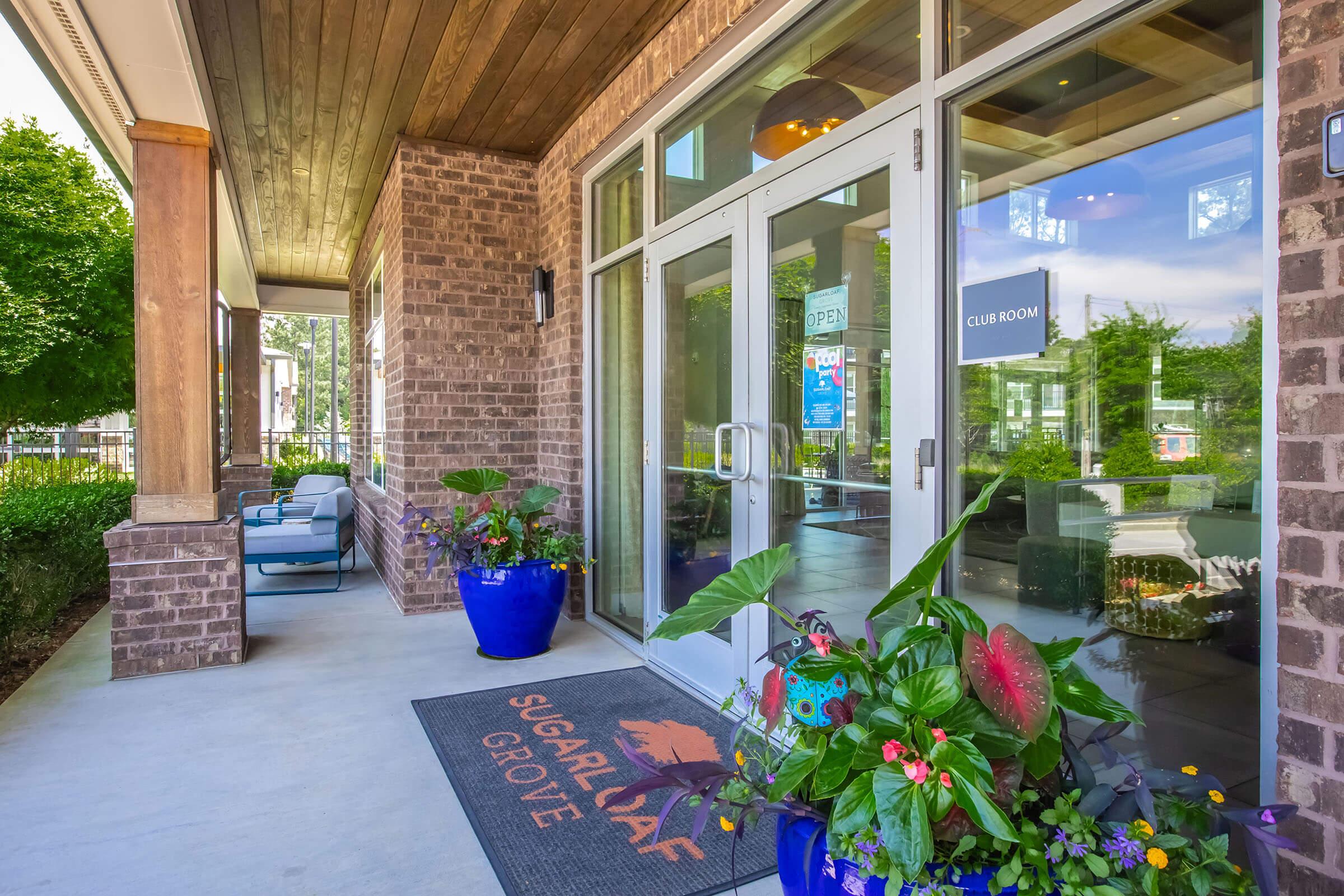 A welcoming entrance to a modern building featuring large glass doors, a "Club Room" sign, and a decorative mat that reads "SUGARLOAF GROVE." The area is adorned with vibrant potted plants and flowers, and there are seating arrangements visible in the background under a shaded porch area.