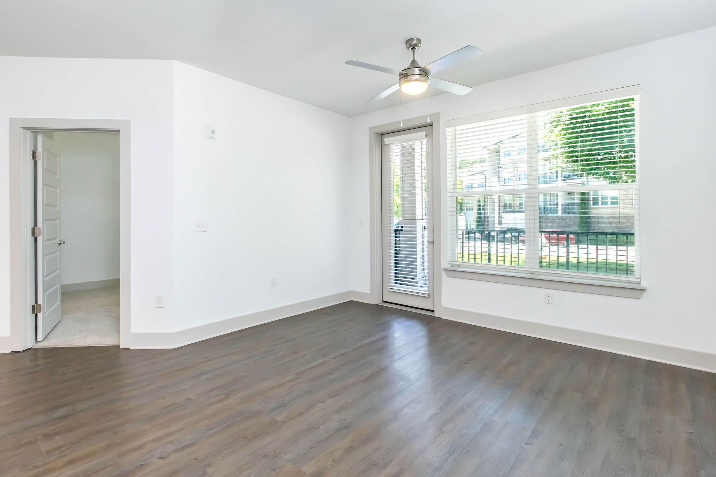 A bright, modern living space featuring light-colored walls, a ceiling fan, and large windows with blinds. The floor is finished in a dark wood look, and a sliding glass door leads to an outdoor area. There is an open doorway to another room on the left.