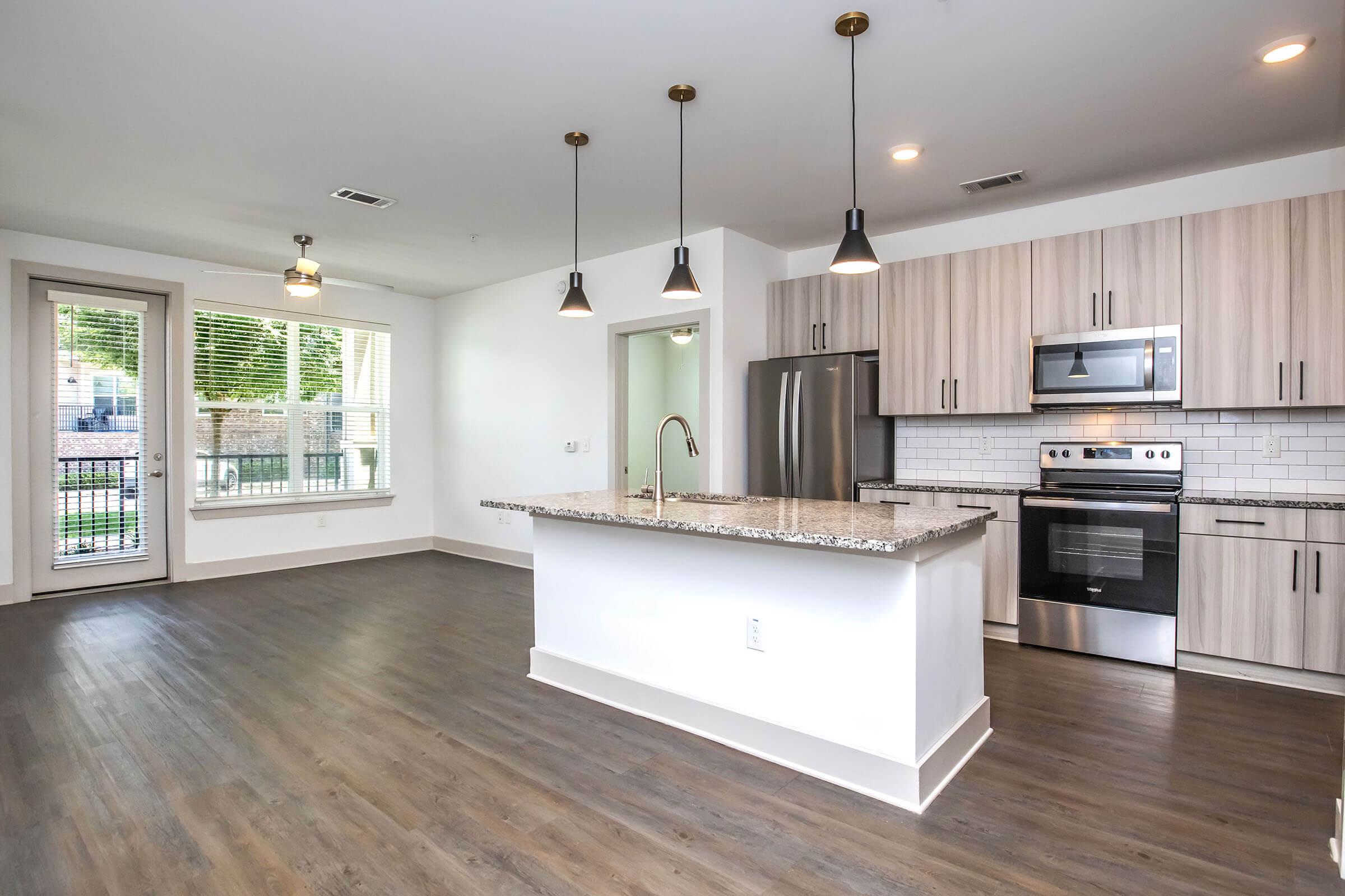 A modern kitchen and living area with a spacious layout. The kitchen features stainless steel appliances, a granite countertop, and pendant lighting. Large windows provide natural light, and there's a sliding glass door leading to an outdoor space. The floor is wooden, adding warmth to the design.
