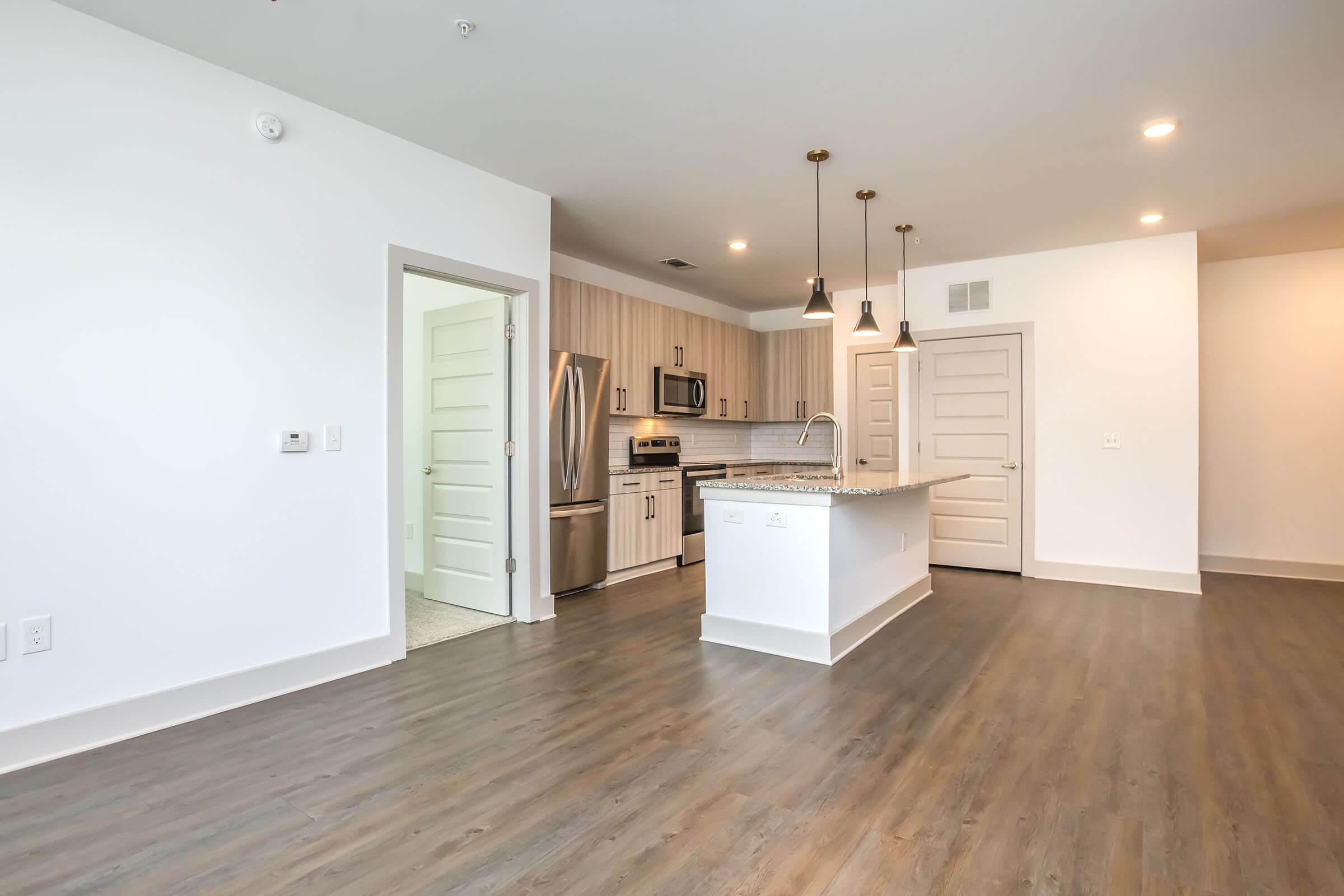 Modern kitchen and living area featuring an open layout. The kitchen includes stainless steel appliances, a large island with seating, and light wood cabinetry. The floor is a stylish laminate, and there are pendant lights hanging above the island. The walls are painted white, enhancing the bright and airy feel of the space.