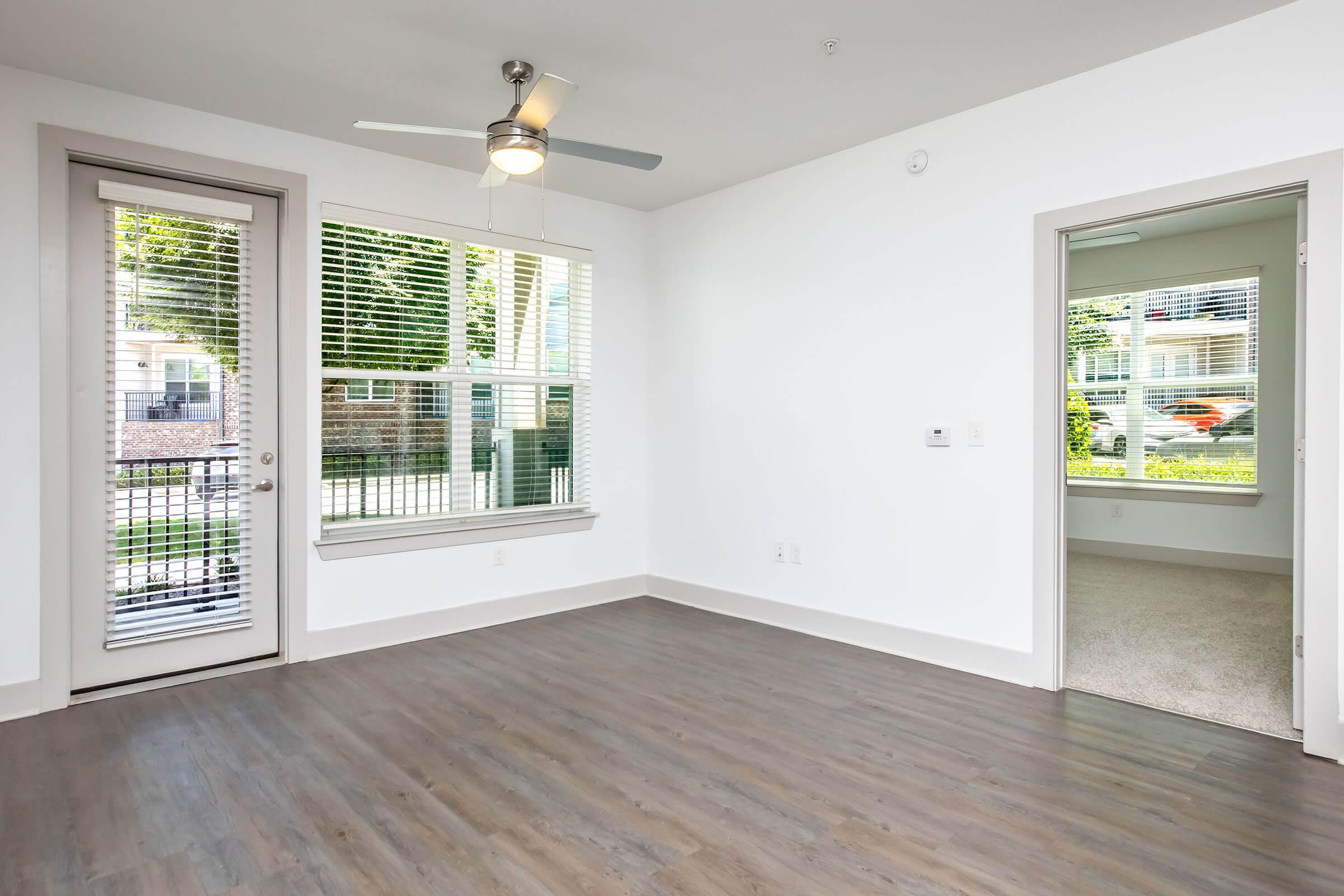 A modern, unfurnished living room featuring a ceiling fan, large windows with blinds, and light-colored walls. One wall has a sliding glass door leading to a balcony, while another door opens to a room with carpet. The flooring is laminate, providing a clean and spacious atmosphere.