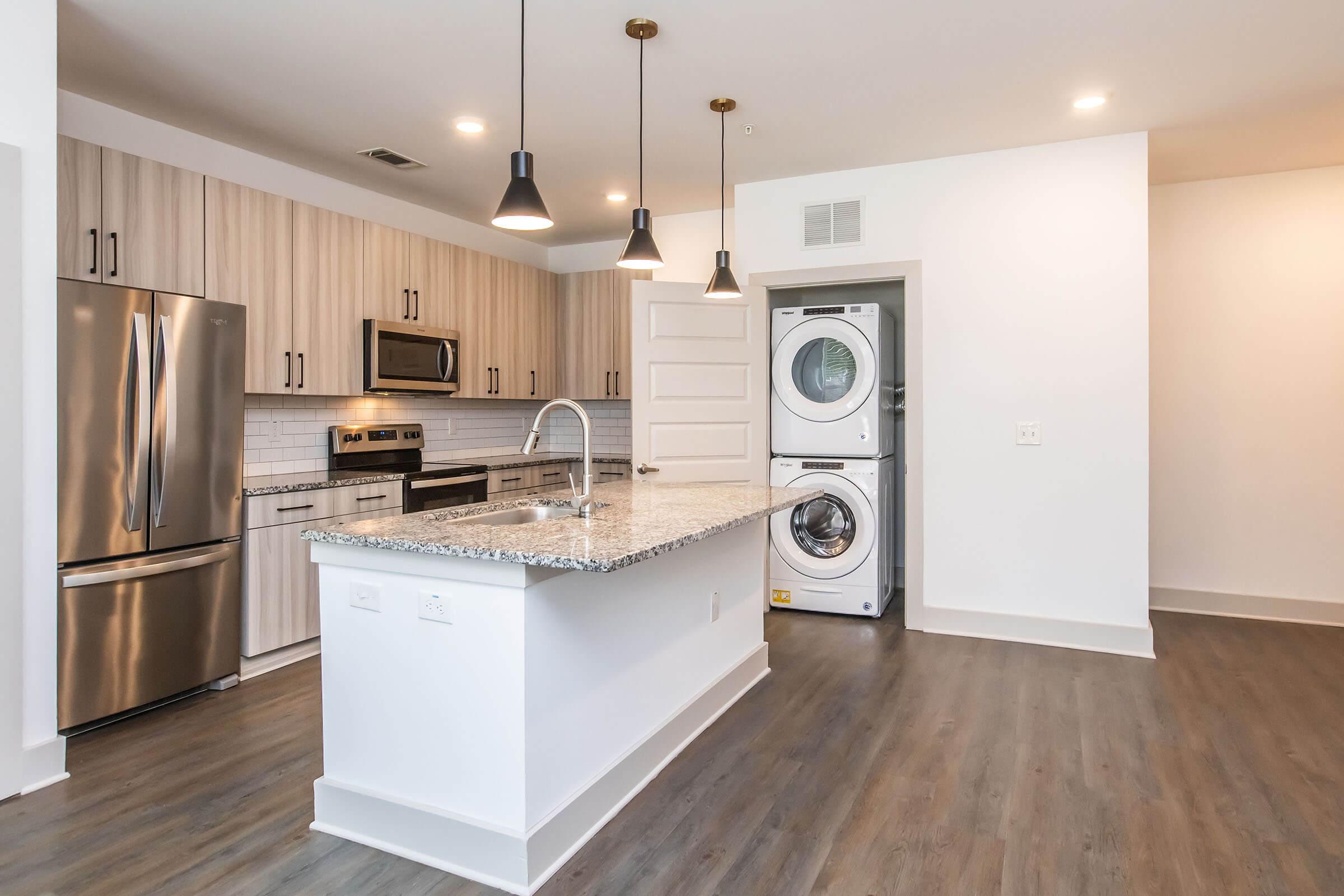 Modern kitchen featuring stainless steel appliances, including a refrigerator and microwave. A granite countertop island with a sink is in the center, and there are pendant lights hanging above. A laundry area with a washer and dryer is visible to the side, and the flooring is a wood-like laminate.