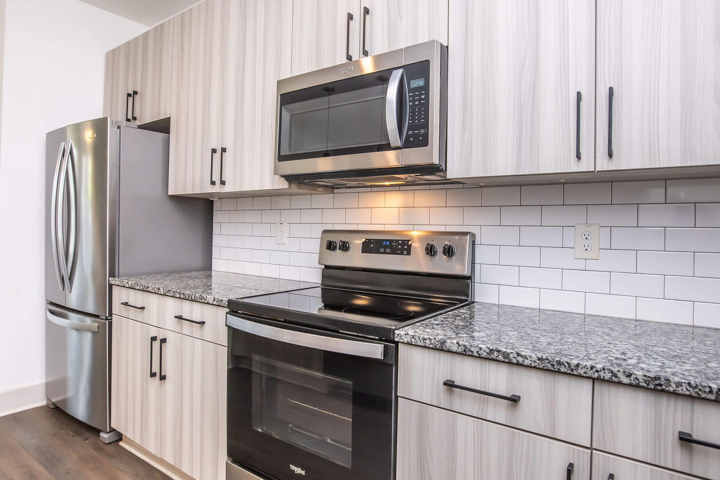 Modern kitchen featuring a stainless steel refrigerator and microwave, a black oven, and white tile backsplash. The cabinets have a light wood finish with black handles, and the countertop is made of dark granite.