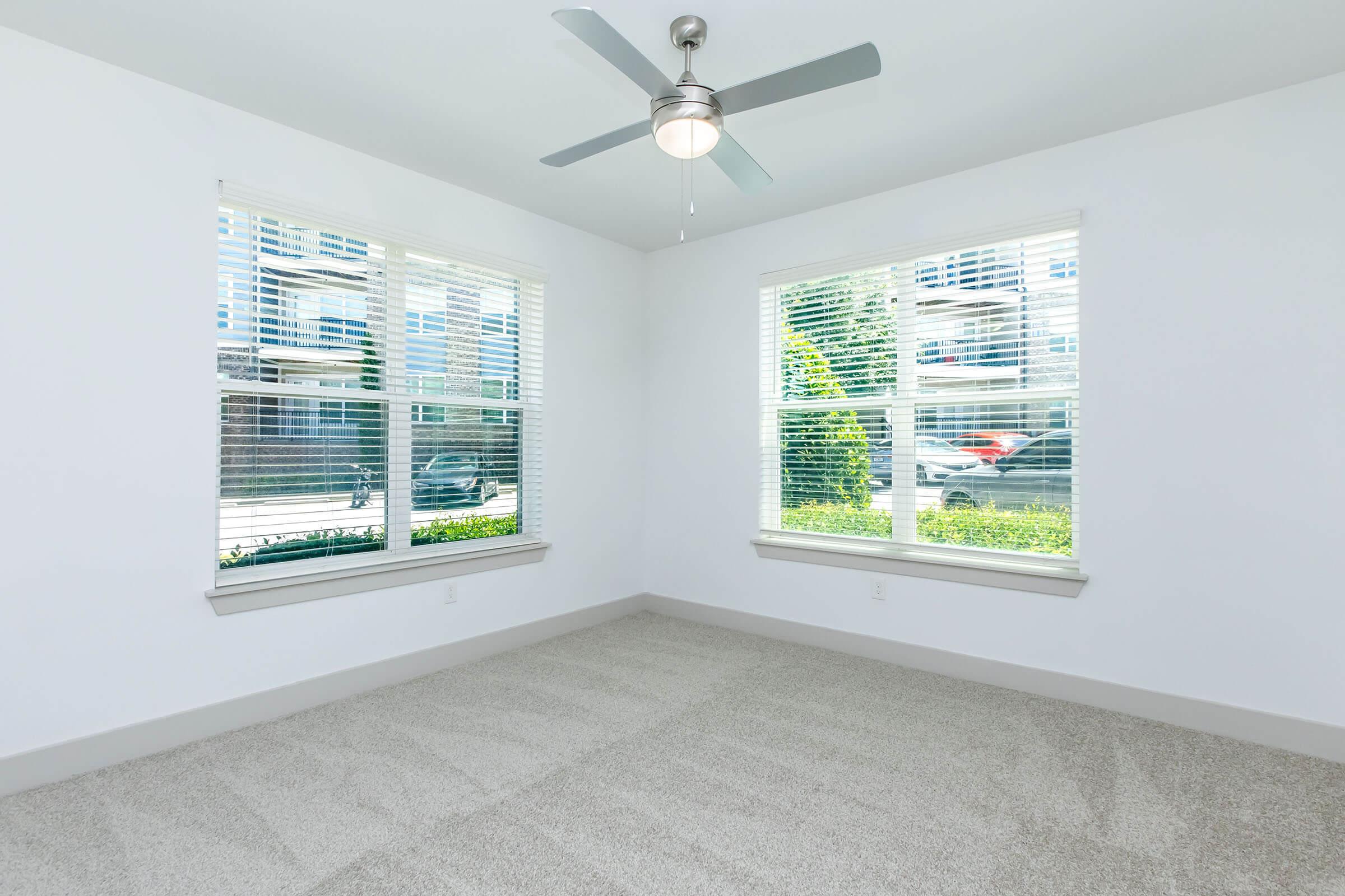 A bright, empty room with light beige carpet and light gray walls. It features two large windows with white blinds, allowing natural light to fill the space. A ceiling fan is mounted in the center, and the room has a modern, clean appearance, ideal for various uses such as an office or living area.