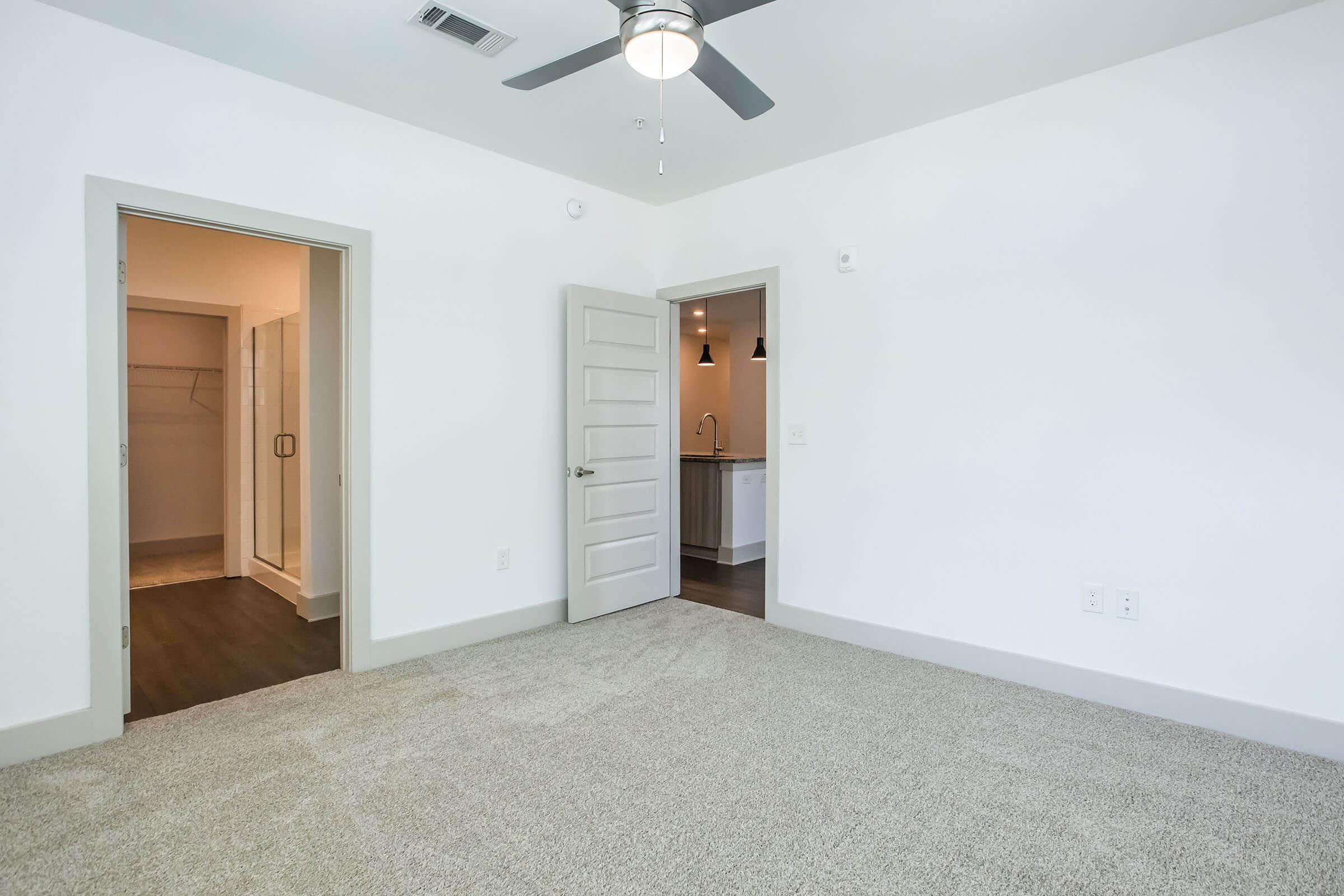Bright, spacious room with light-colored walls and plush carpet. Two doorways lead to a closet and a kitchenette area. A ceiling fan is mounted in the center of the room, enhancing airflow. The overall aesthetic is modern and inviting, suitable for a cozy living space.
