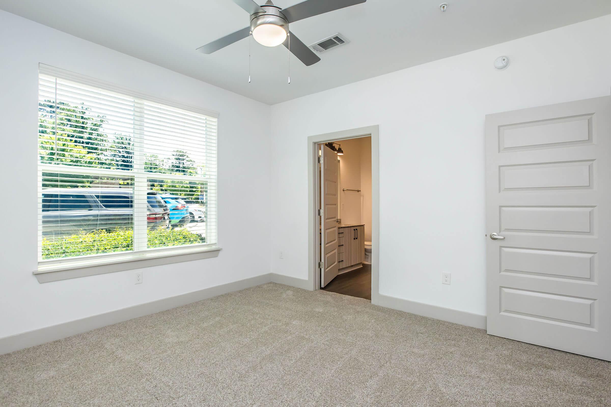 A bright, empty room with light gray carpet, a ceiling fan, and a window featuring white blinds. The door leading to another room is open, revealing a small area with light-colored cabinets. Natural light illuminates the space, enhancing the feeling of openness.