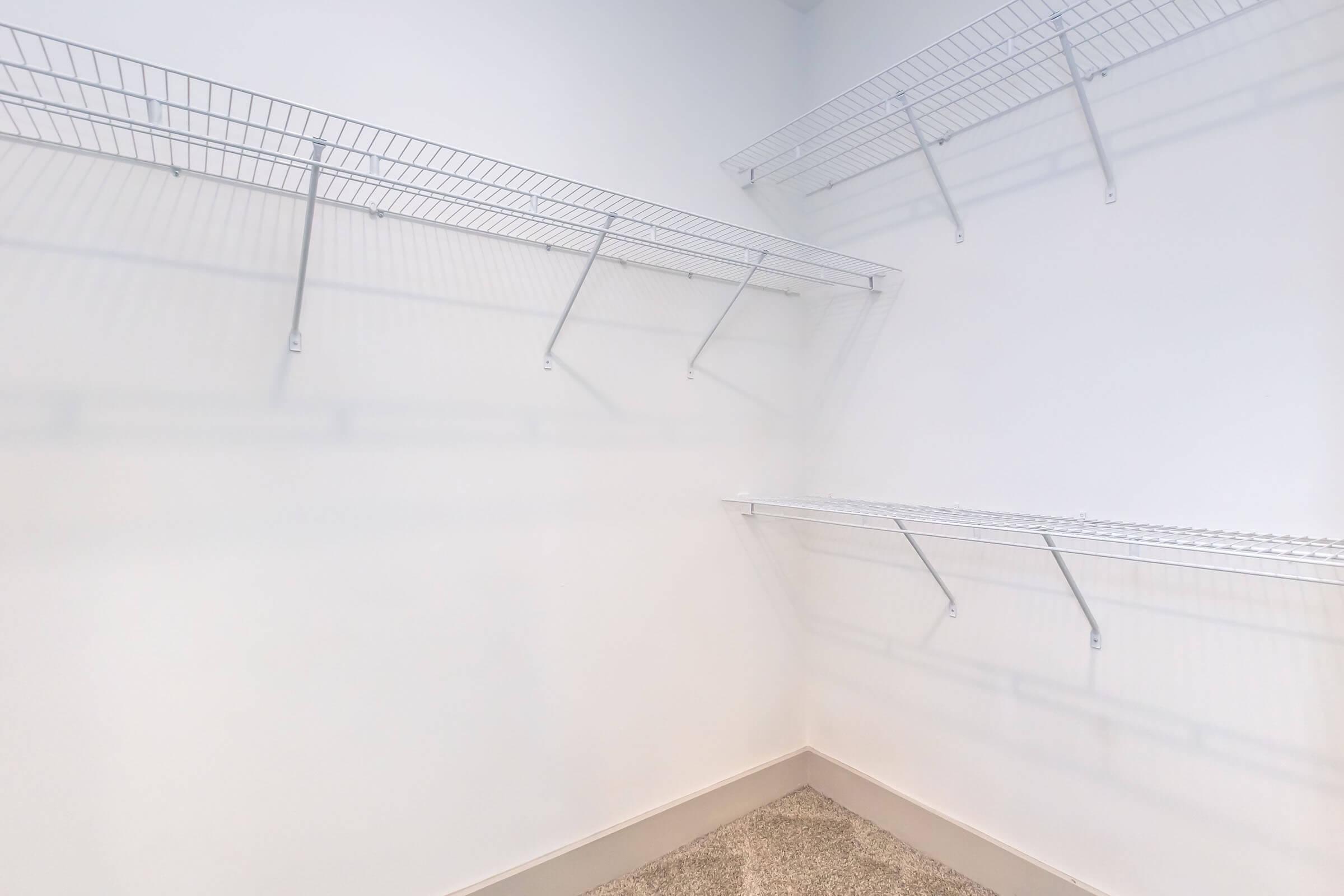 Empty closet with white wire shelving mounted on the walls. The walls are painted light, and the floor is covered in beige carpet. The shelving units are arranged in a way that creates open space, emphasizing the minimalistic design.