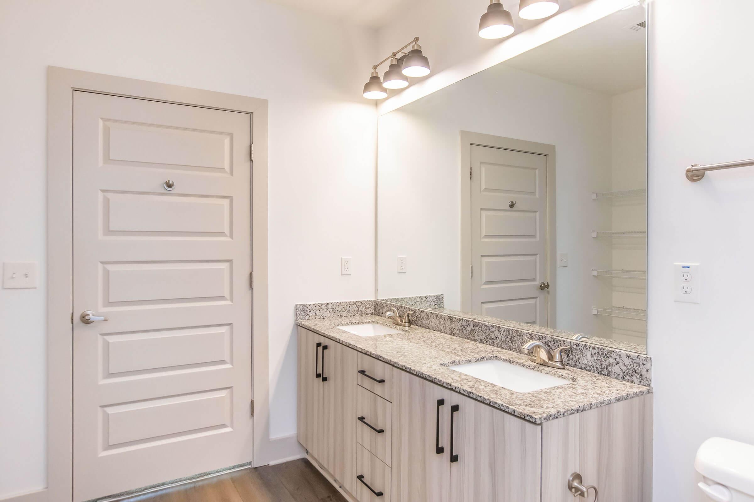 A modern bathroom featuring a double vanity with granite countertops, two sinks, and stylish faucet fixtures. A large mirror spans the wall above the vanity, reflecting light from three overhead lights. The room includes a white door and built-in shelving on the opposite wall. The flooring is a warm wood tone.