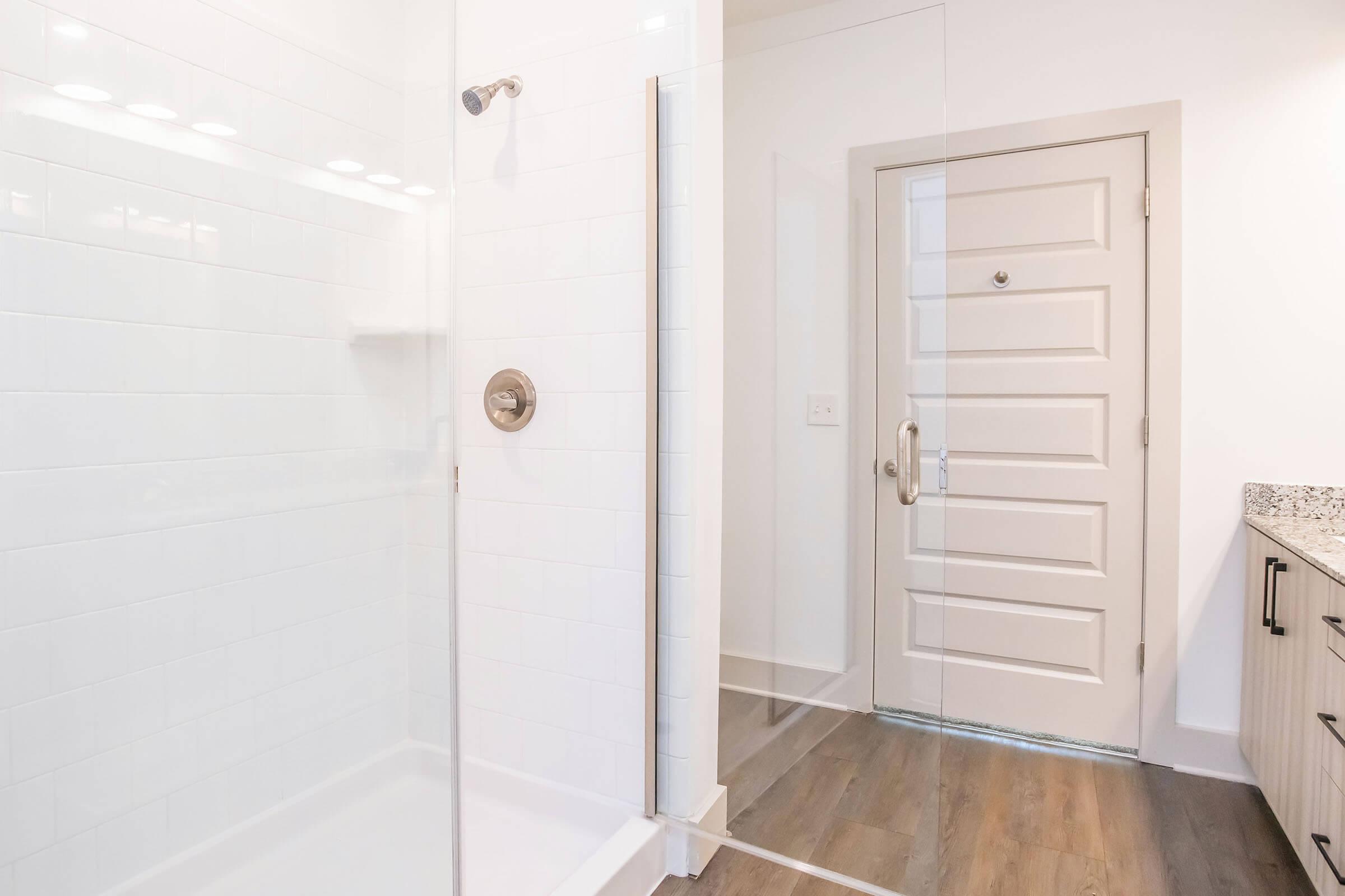 Modern bathroom featuring a glass shower enclosure, white tiled walls, and a granite countertop. The floor has a wood-like finish, and there's a door leading to another room, indicating a contemporary design. Bright lighting enhances the spacious and clean appearance of the area.