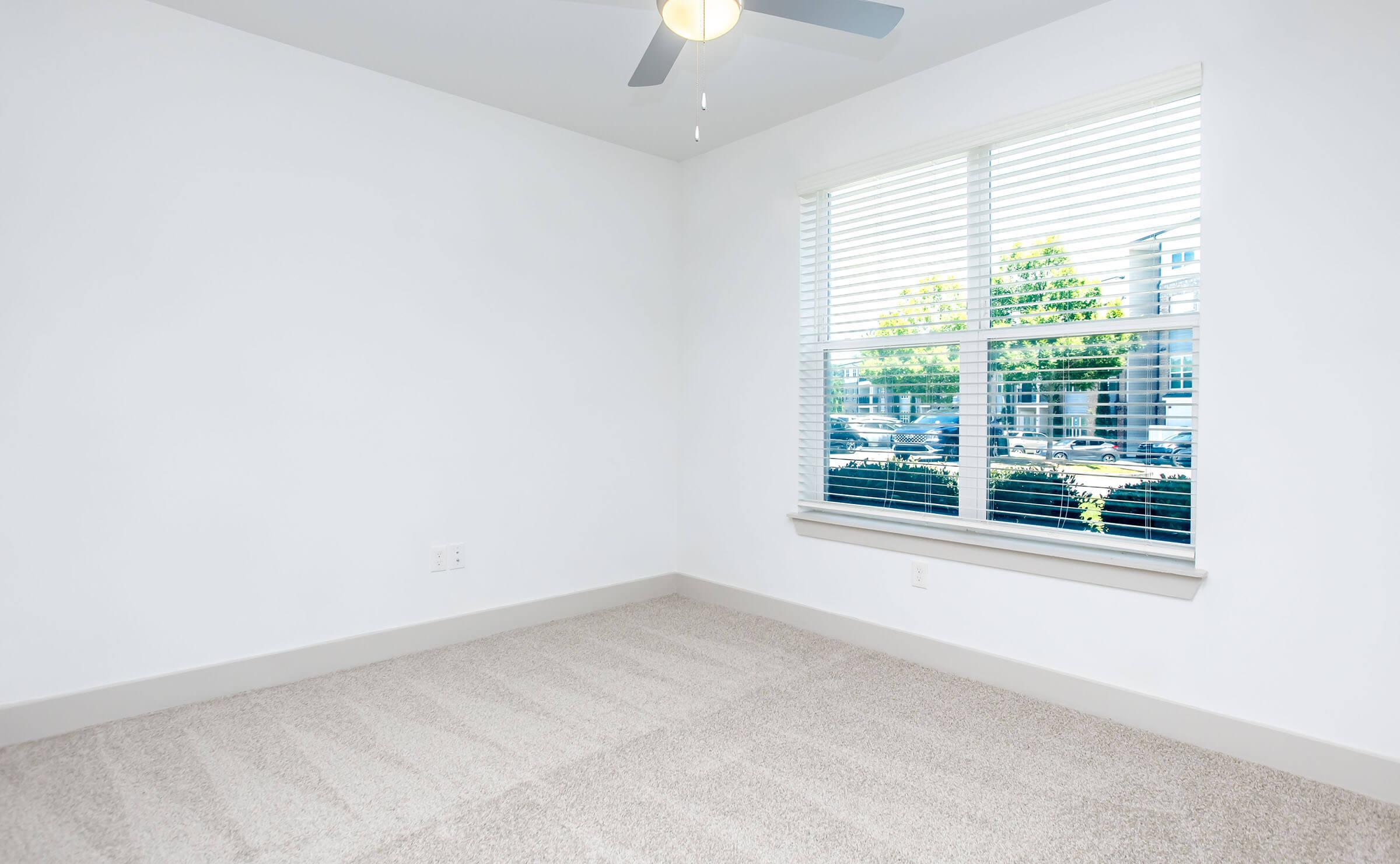 Empty room featuring light-colored walls and a soft carpet. A ceiling fan hangs from the ceiling, and a large window with white blinds allows natural light to illuminate the space, providing a view of greenery outside. The room appears modern and well-maintained.