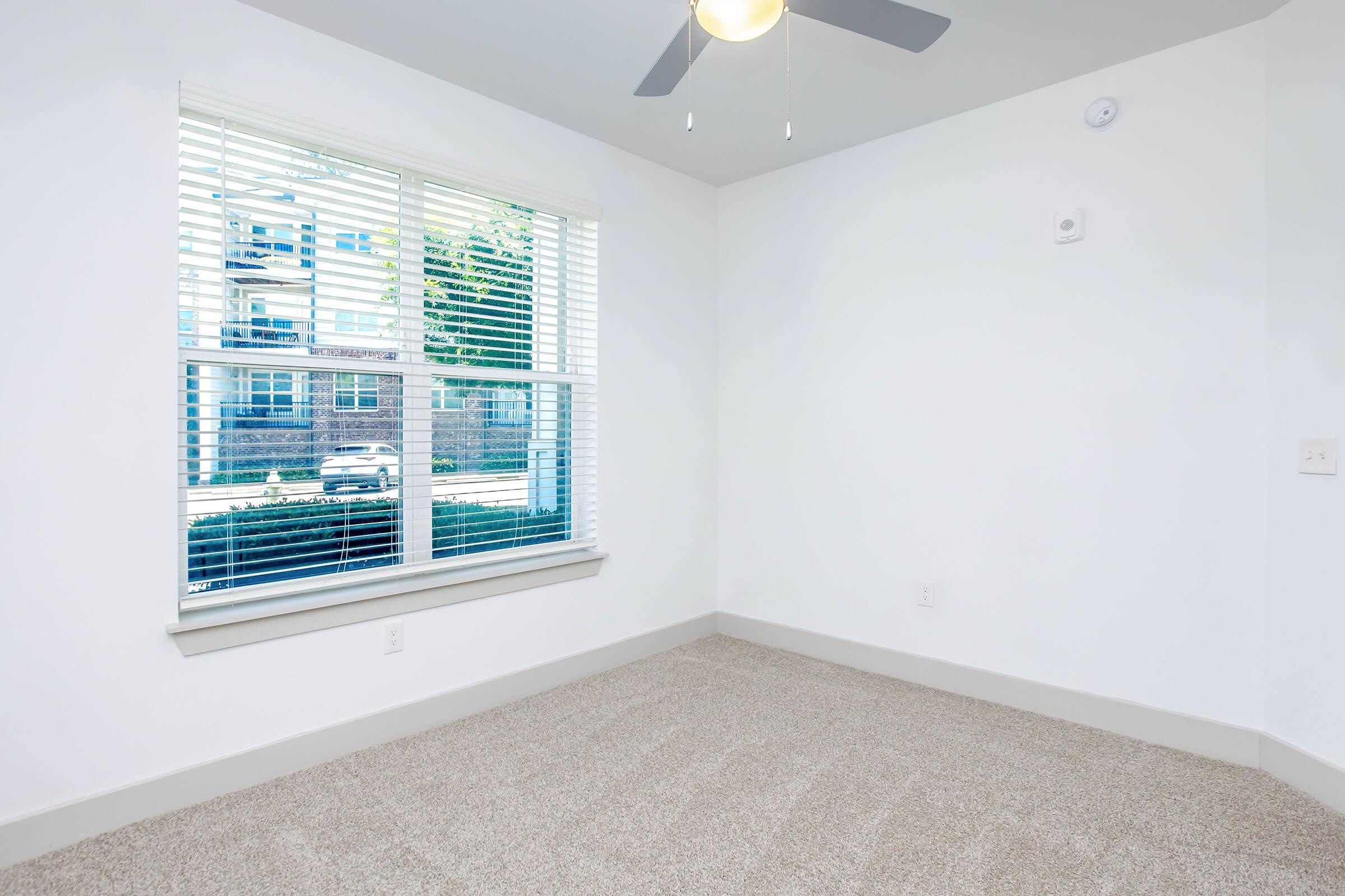 A bright, empty room with light-colored walls and a ceiling fan. A large window with white blinds offers a view of outside, revealing a clear view of another building and street. The floor is covered in beige carpet, creating a clean and inviting atmosphere.
