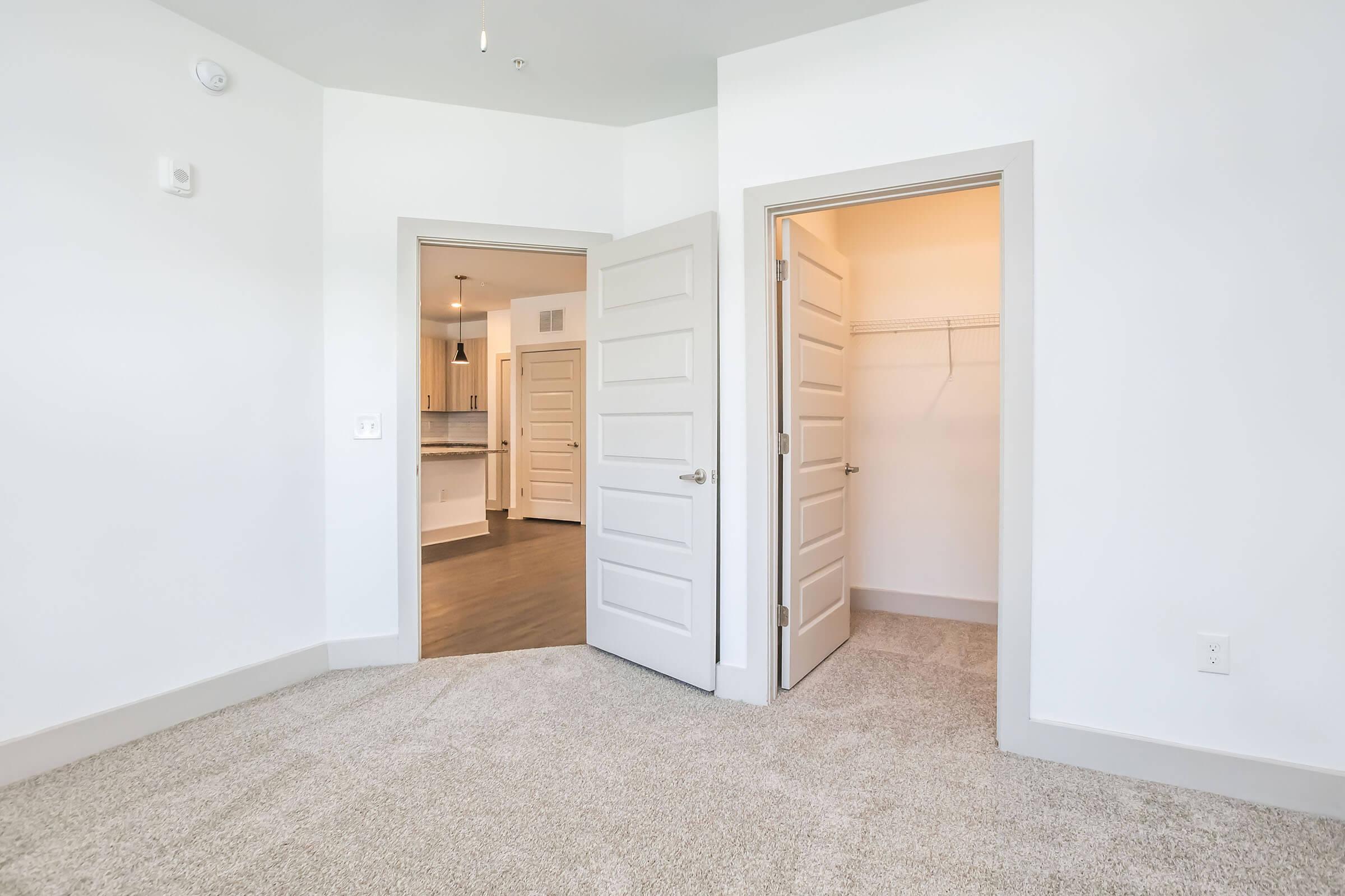 Interior view of a room with two open doors leading to separate areas. The room has light-colored walls and a carpeted floor. One door opens to a closet with a hanging rail, while the other leads to a kitchen or living area visible in the background. Natural light brightens the space.