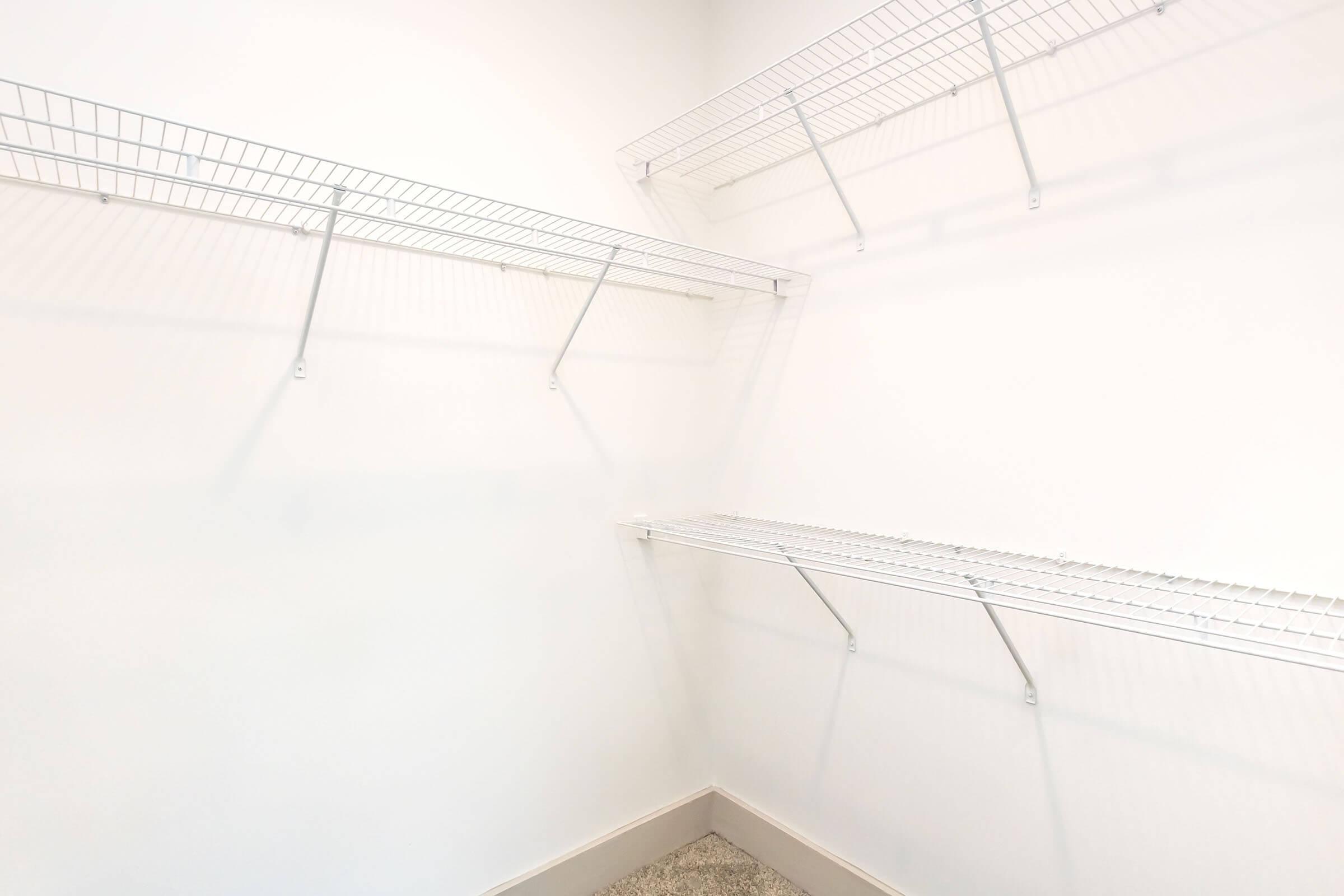 A clean, empty closet featuring white wire shelving mounted on light-colored walls. The shelves are arranged on three sides, creating an organized and spacious storage area. The floor is carpeted, adding a touch of warmth to the minimalist design.