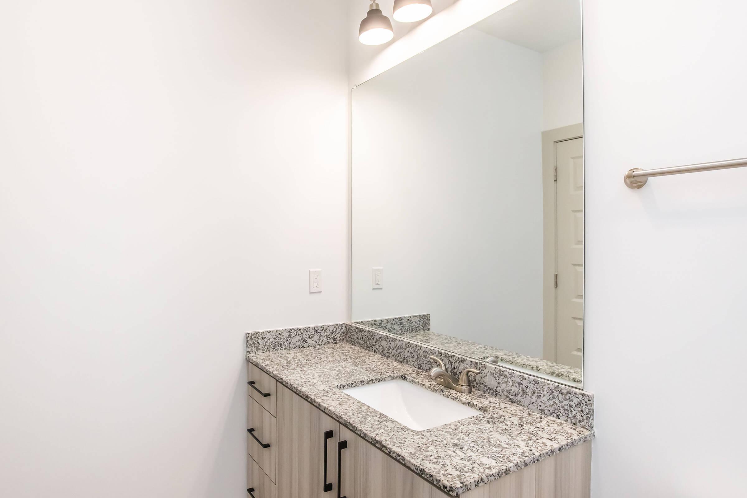 A modern bathroom featuring a sleek granite countertop with an integrated sink, a large wall mirror above, and minimalist cabinetry. The space is well-lit by two pendant lights, and the walls are painted white, creating a clean and spacious atmosphere.