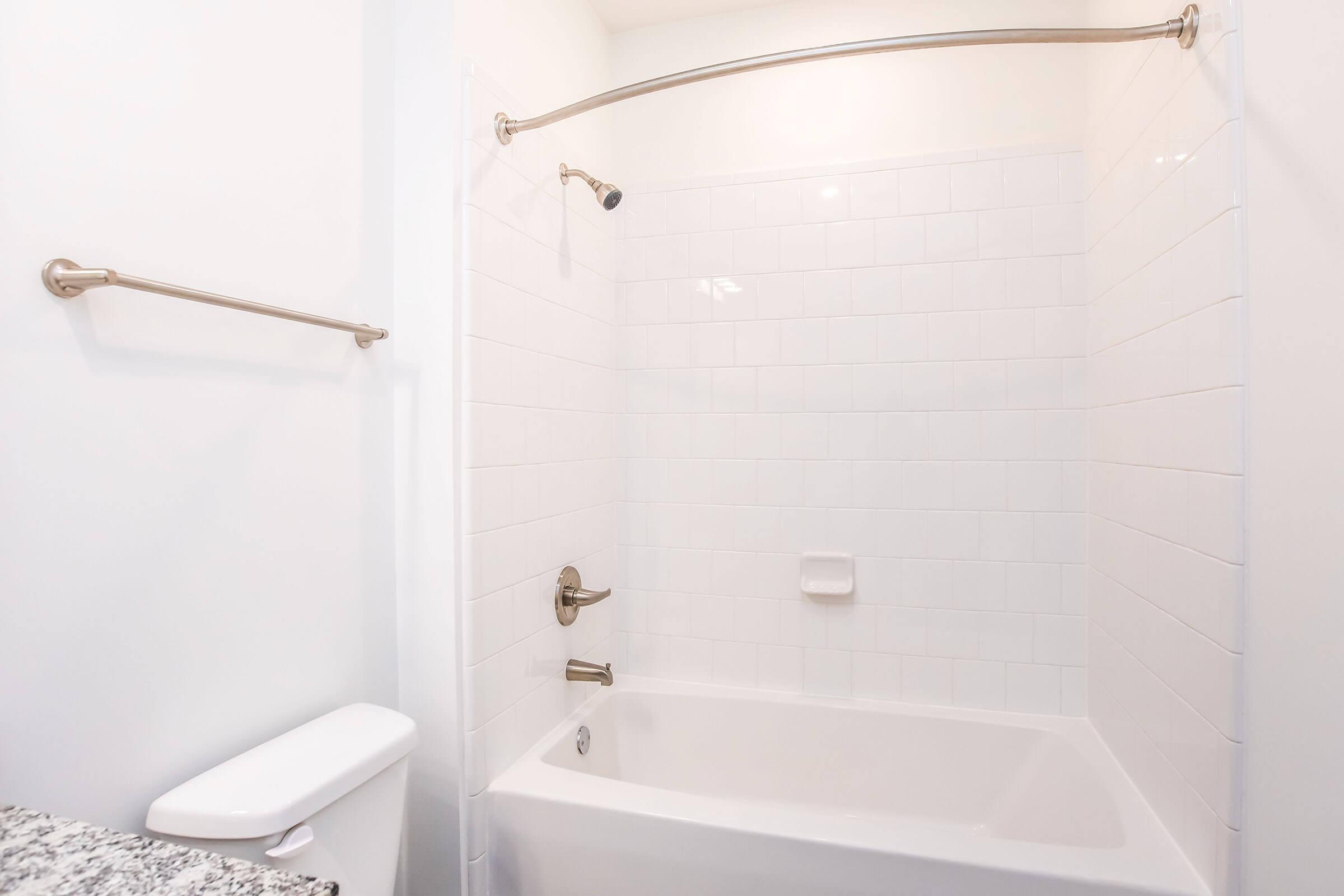 A clean bathroom with a white tiled shower and bathtub. The shower is equipped with a modern faucet and a curved shower rod. A small shelf is visible on the wall and there’s a toilet in the corner next to a granite countertop, creating a spacious and bright atmosphere.