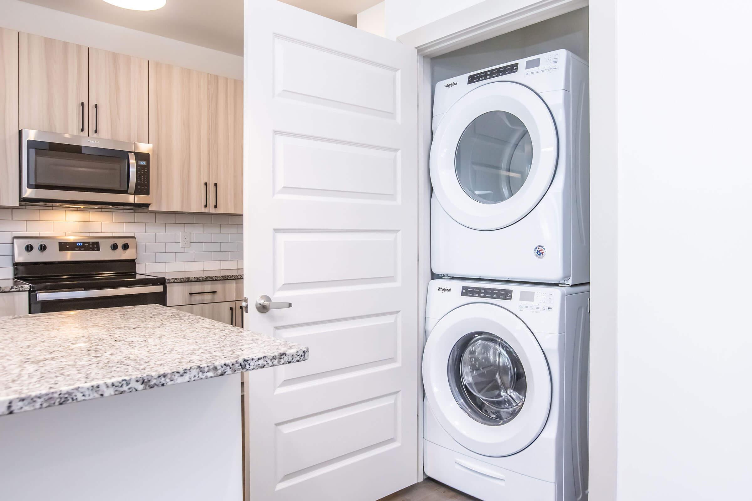 A modern kitchen featuring light wood cabinetry, a stainless steel microwave and oven, and a granite countertop. In the foreground, a door is open, revealing a stacked washer and dryer in a small closet space. The kitchen has a clean, contemporary aesthetic with bright lighting.