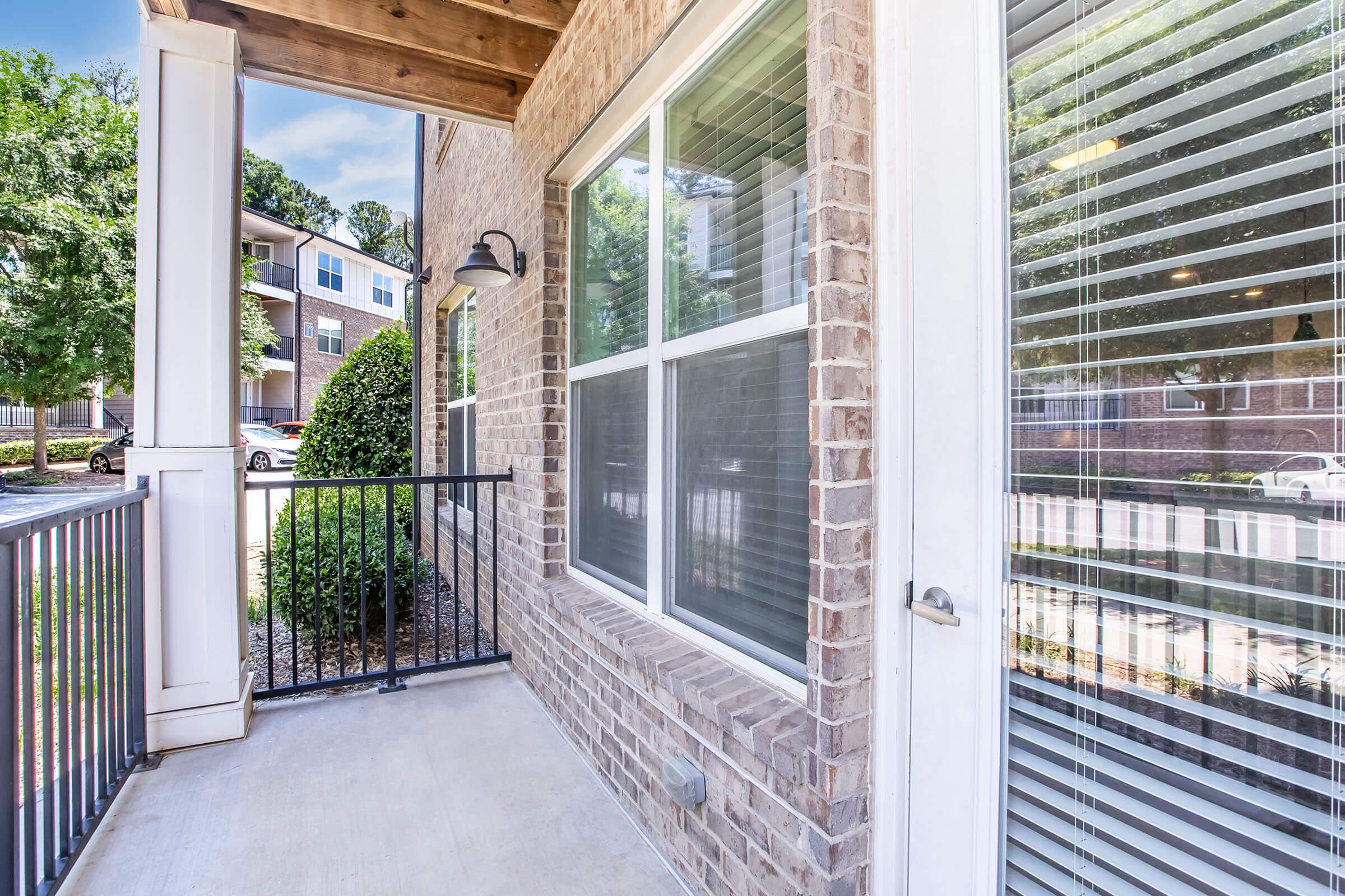 A corner balcony view featuring brick walls and a railing, with large windows allowing natural light. Lush green bushes are outside, and the scene includes nearby apartment buildings and a clear blue sky. The area is well-lit and inviting.