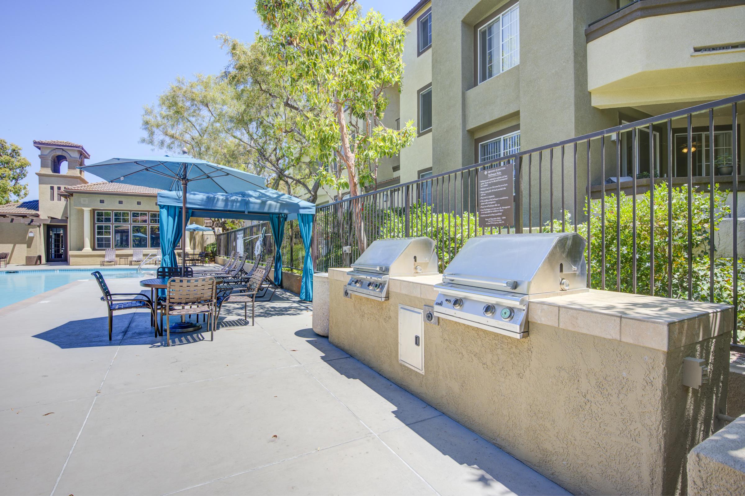 Outdoor grilling area beside a swimming pool, featuring two stainless steel barbecue grills, a shaded seating area with a blue umbrella, and a row of chairs and tables. Lush greenery surrounds the area, with a residential building in the background, creating a relaxing atmosphere for gatherings.