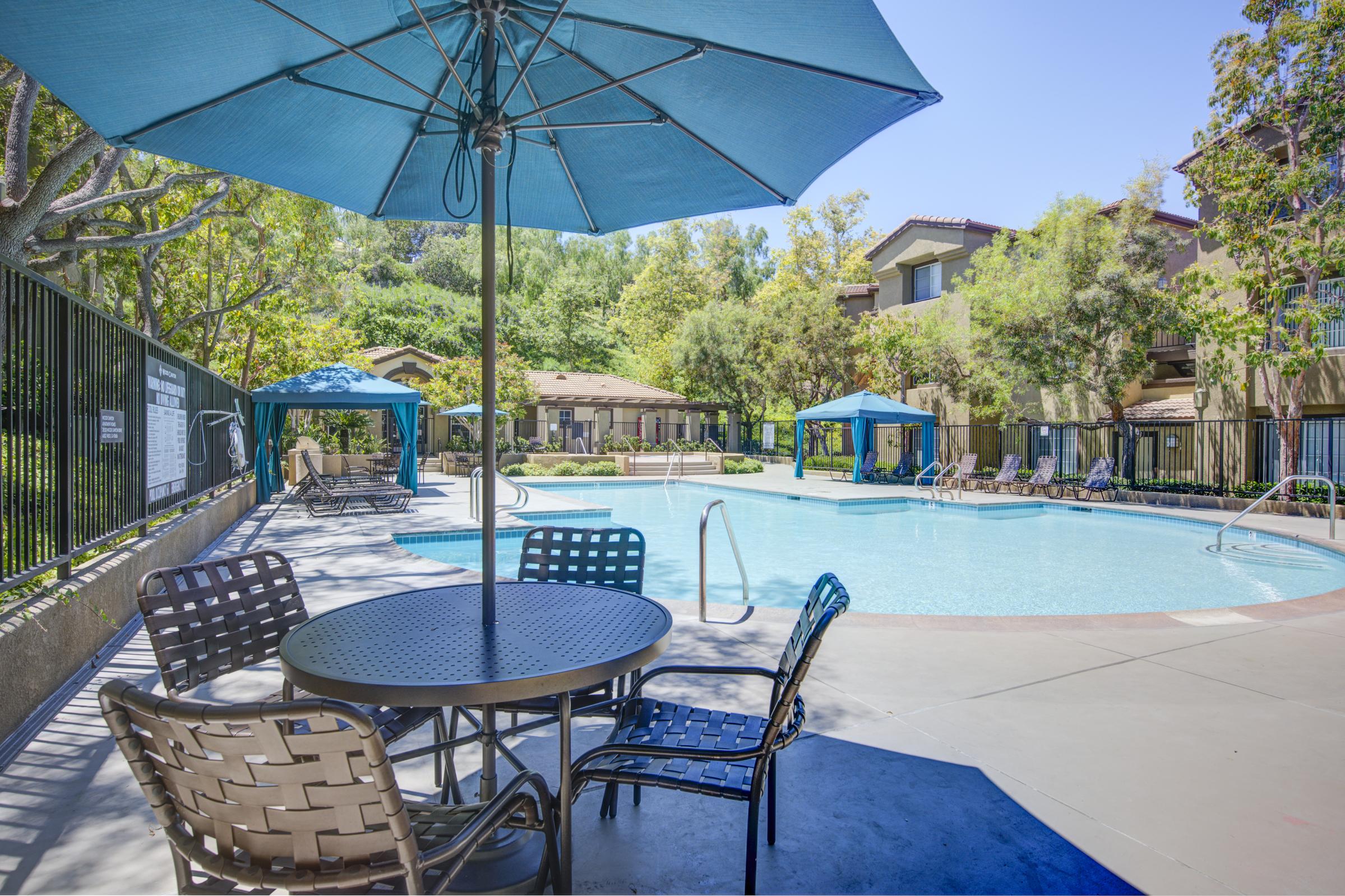 An outdoor swimming pool surrounded by lounge chairs and umbrellas. A shaded table with chairs is in the foreground, while trees and residential buildings are visible in the background under a clear blue sky.