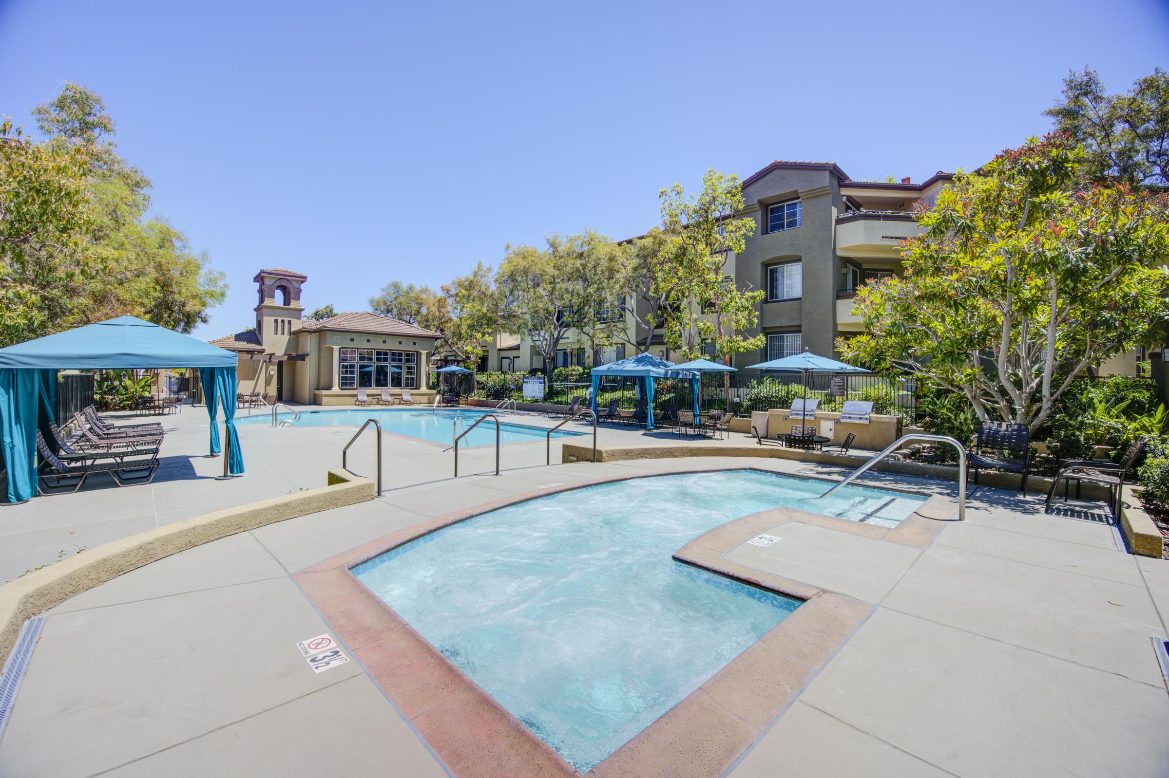 A sunny outdoor pool area featuring a large swimming pool, a hot tub, and surrounding lounge chairs. Several blue shaded cabanas are positioned nearby, with greenery adding to the tranquil atmosphere. Modern apartment buildings are visible in the background under a clear blue sky.