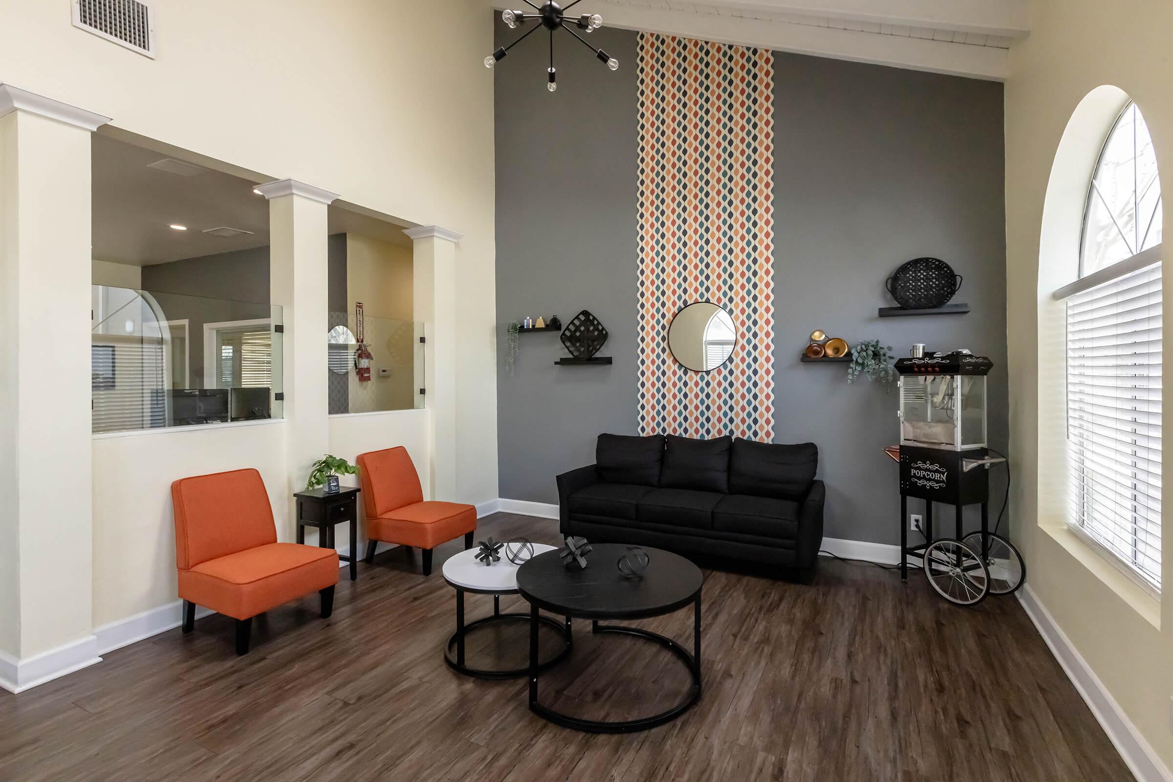 A cozy lounge area featuring a black sofa, two orange chairs, and a circular coffee table. The wall behind is decorated with a geometric pattern and a round mirror. A popcorn machine stands to one side, and there are decorative shelves with various items. Large windows provide natural light.