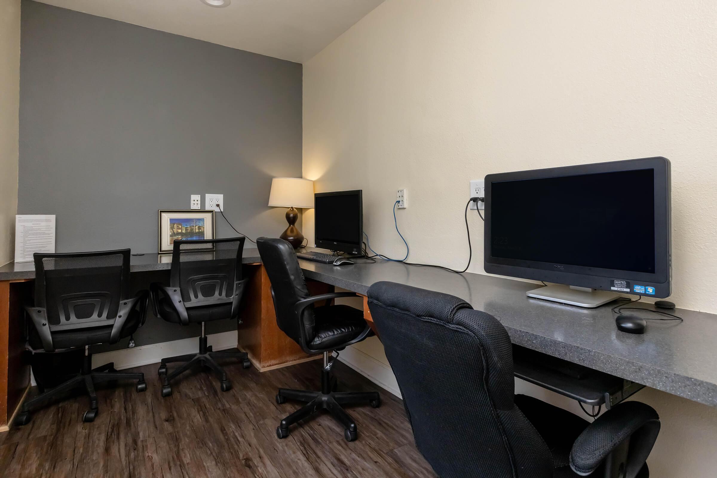 A workspace featuring two computer desks with black office chairs. One desk has a lamp and a framed picture on the wall, while both desks have computer monitors. The walls are painted in neutral tones, and the floor is finished with dark wood-like flooring.