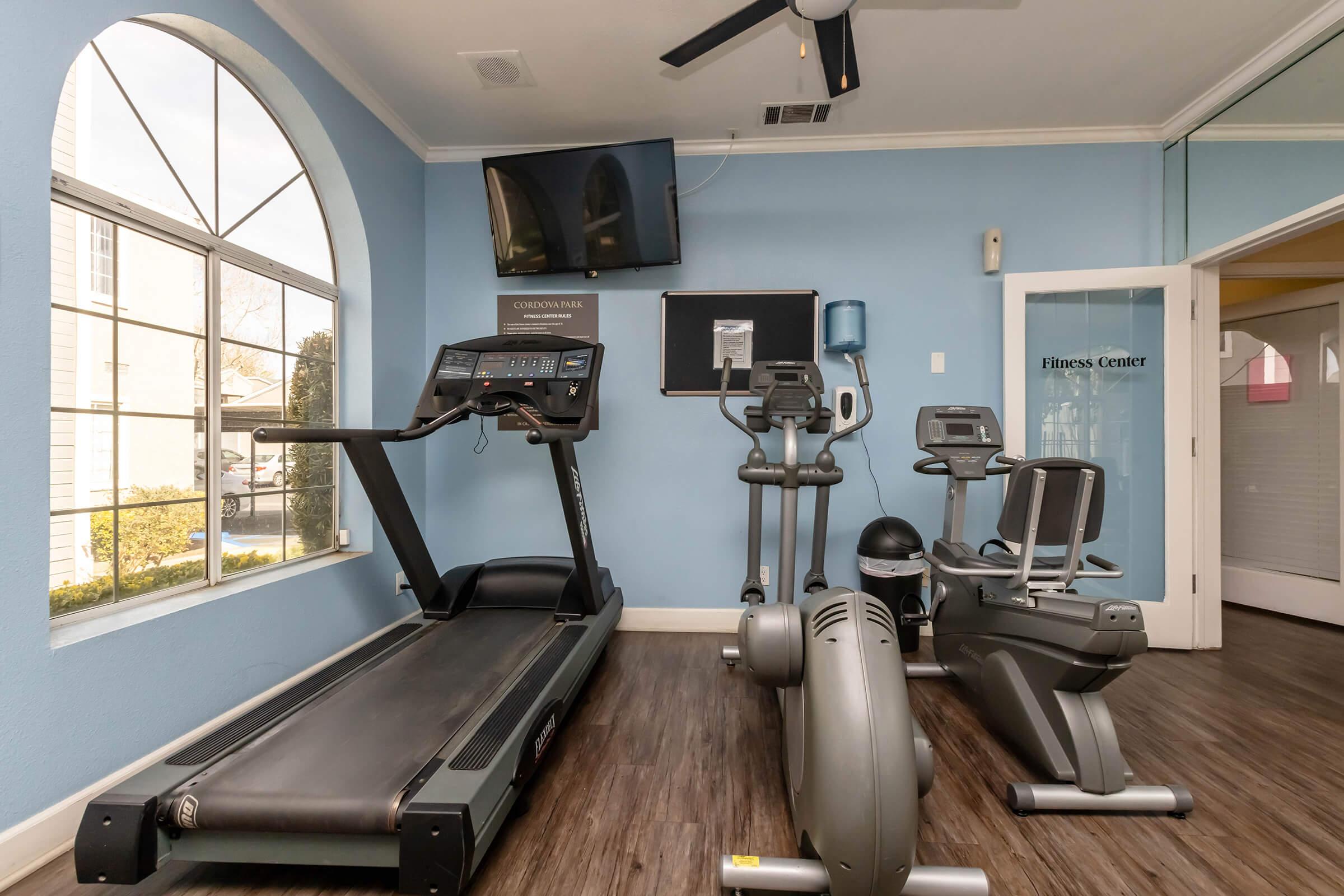 A bright fitness center featuring a treadmill, stationary bike, elliptical machine, and a wall-mounted TV. Large windows allow natural light to illuminate the room, and there's a bulletin board for notifications. The floor has a wooden appearance, creating a warm and inviting atmosphere.