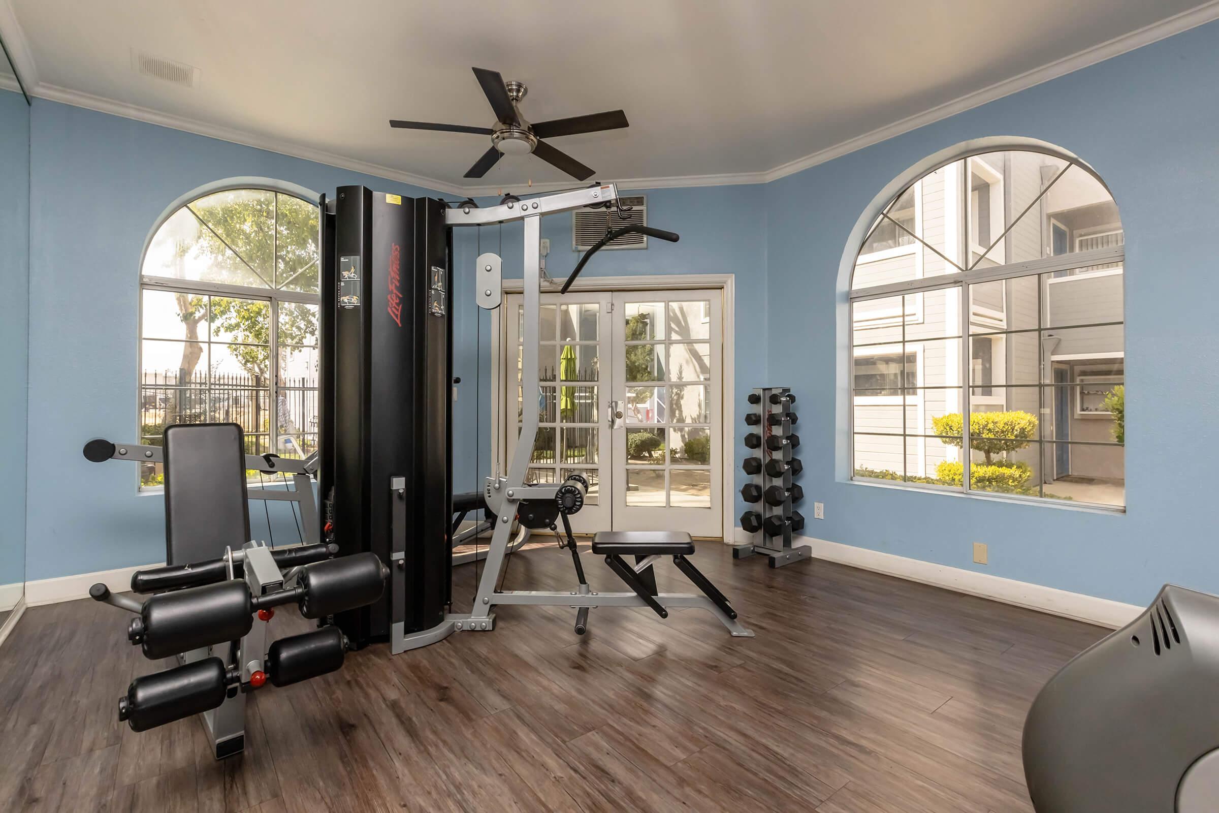 A well-lit gym interior featuring a multi-station workout machine, a weight bench, and free weights. Large windows provide natural light and a view of greenery outside. The room has a modern design with wooden flooring and light blue walls, creating an open and inviting atmosphere for exercise.
