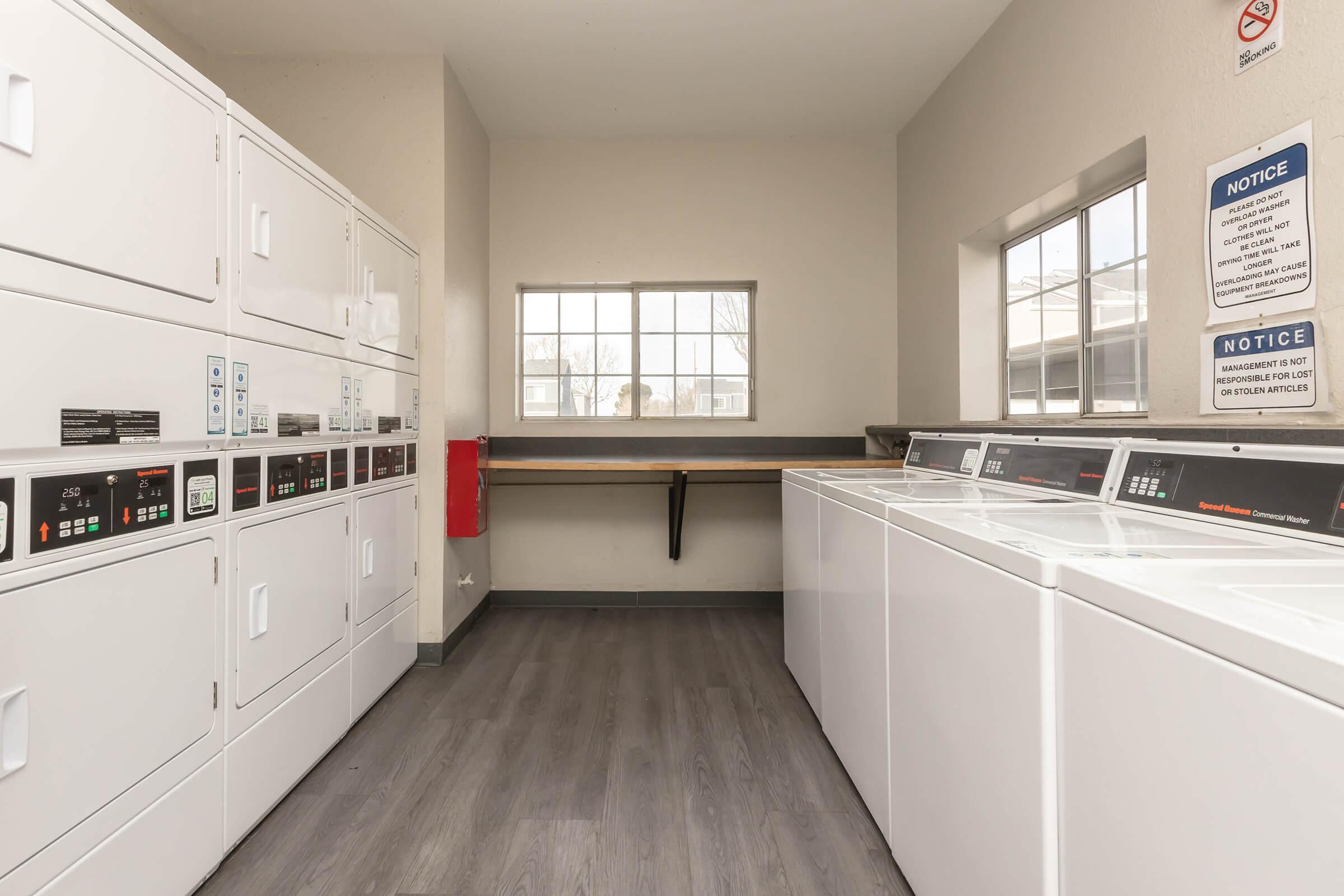 A laundry room featuring rows of white washing machines and dryers along two walls. A window allows natural light to enter, and there's a countertop space for folding clothes. Notices are posted on the walls, and the floor is covered with gray flooring, enhancing the clean and functional atmosphere.