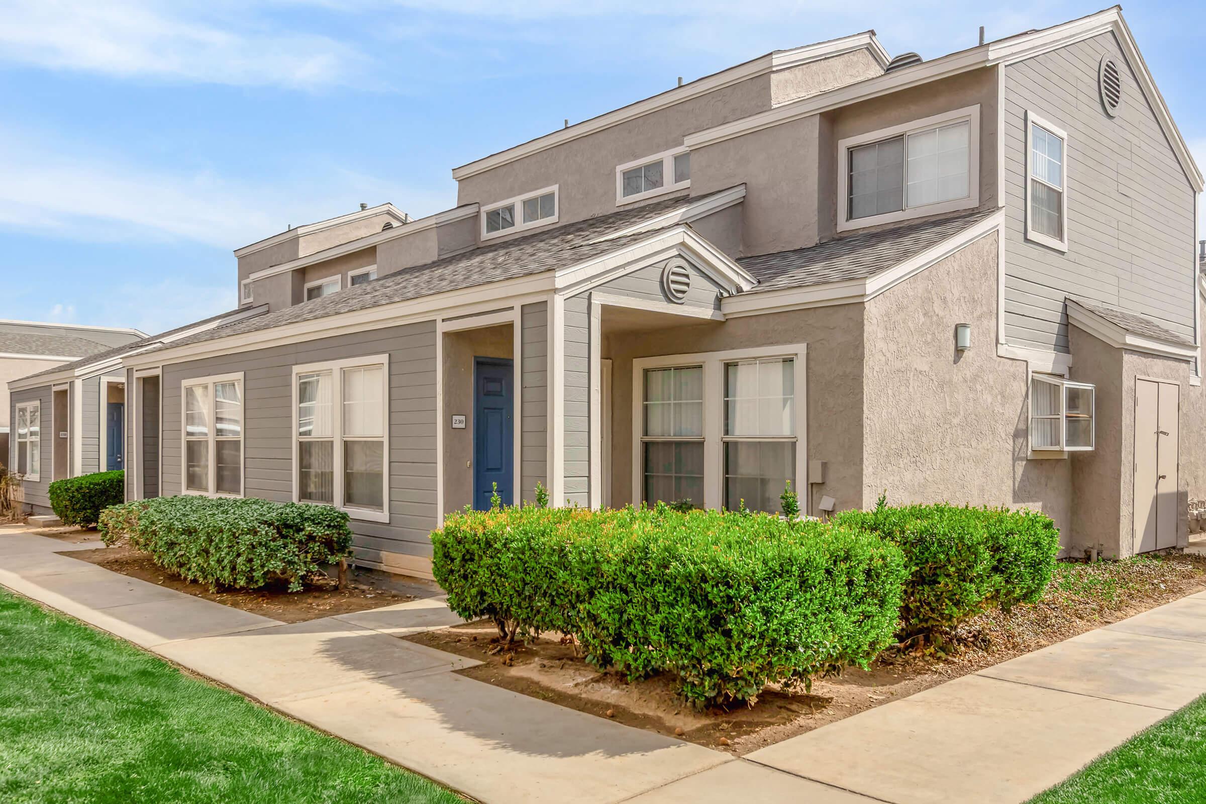 A modern multi-unit residential building with grey siding and a blue front door. The building features a landscaped area with neatly trimmed shrubs and grass, along with a pathway leading to the entrance. Clear blue sky in the background enhances the inviting atmosphere.
