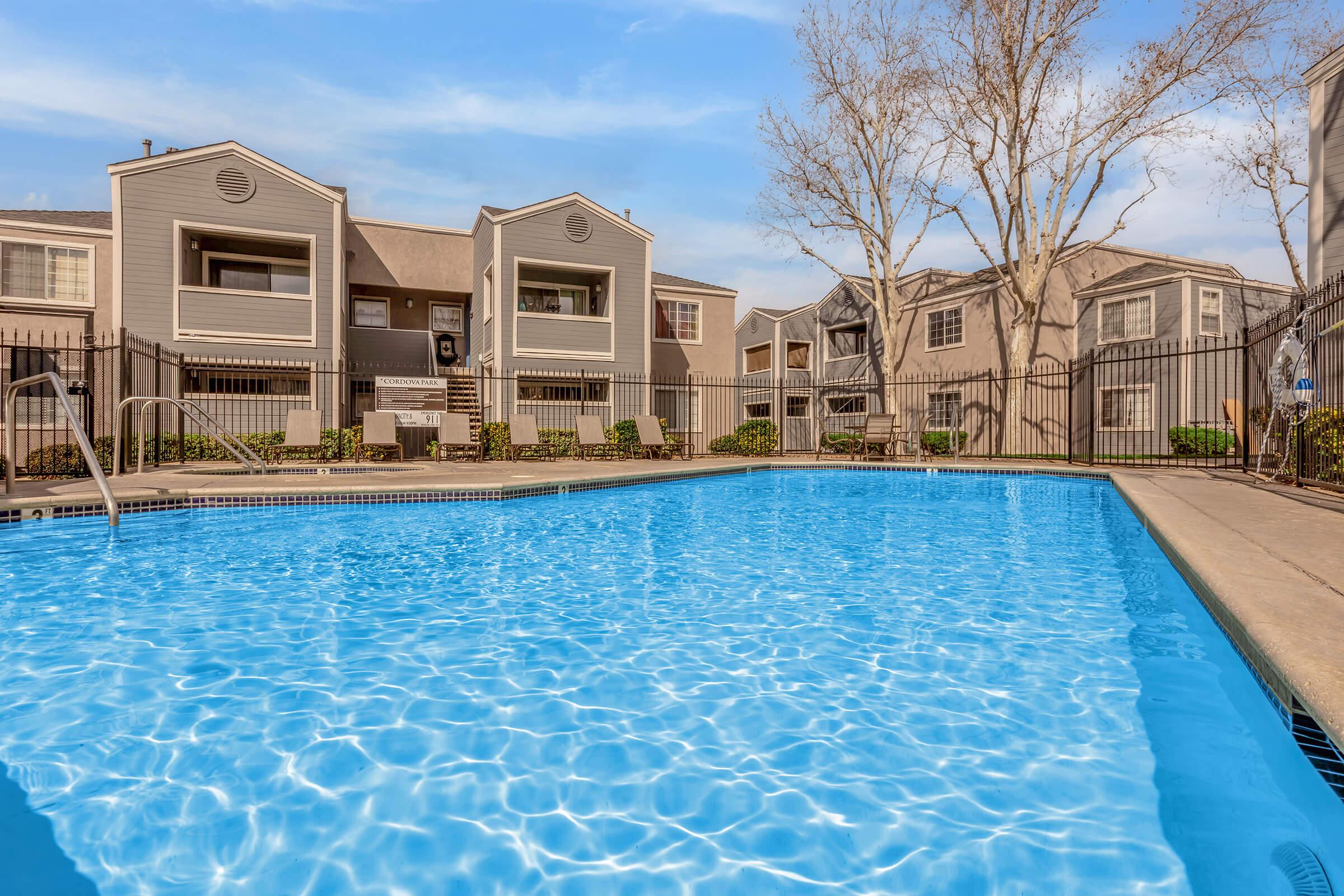 A clear blue swimming pool is in the foreground, surrounded by a fence. In the background, there are two-story apartment buildings with balconies and landscaped areas. Trees are visible nearby, and the sky is bright with some clouds. The scene conveys a relaxing outdoor environment.