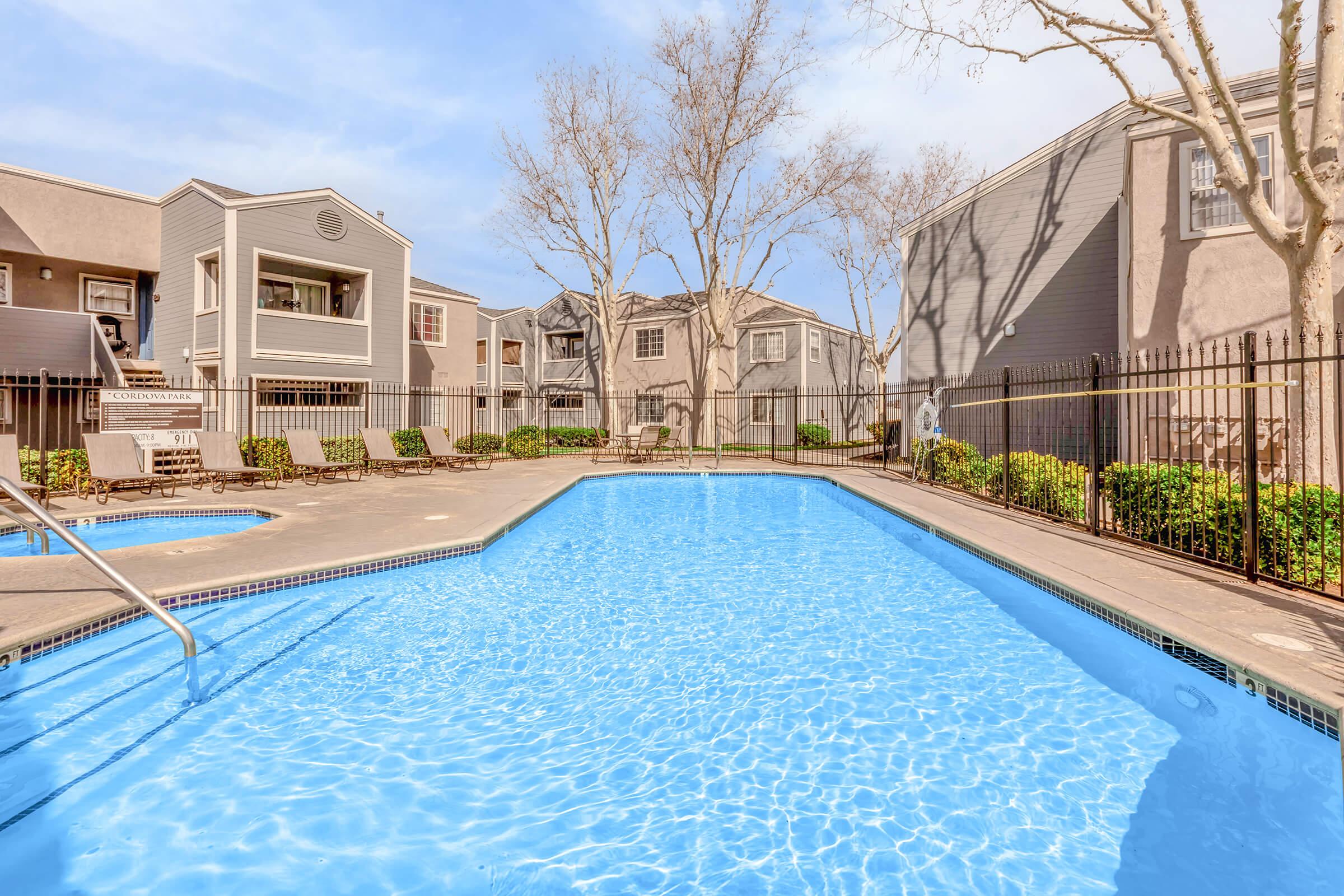 A clear swimming pool surrounded by a fenced area, with lounge chairs and trees nearby. In the background, there are residential buildings with balconies and windows, set in a sunny environment. The water reflects the blue sky, creating a serene atmosphere.